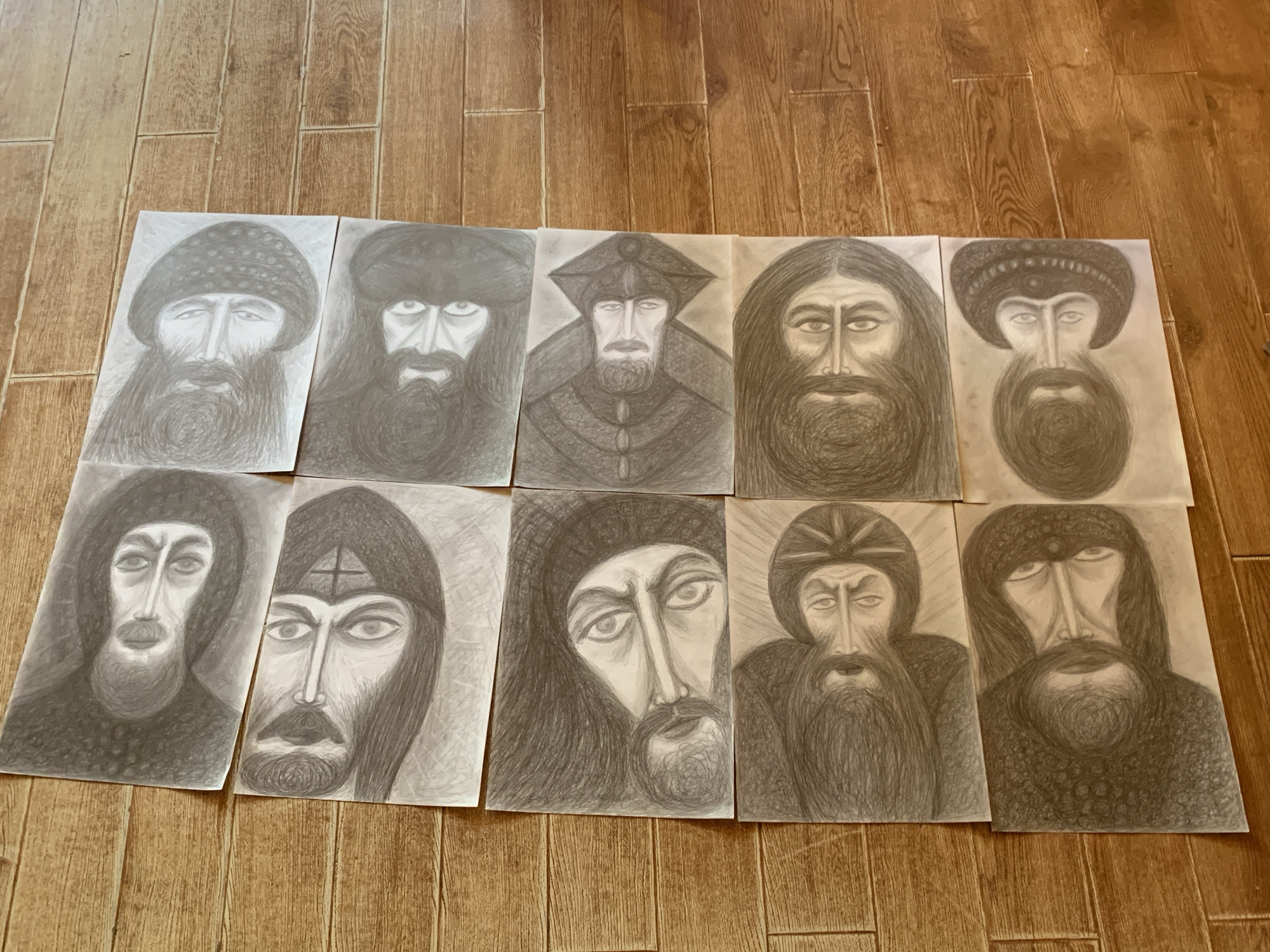 Ten graphite pencil drawings of faces are arranged in two columns on a light brown wooden floor. The drawings are oriented vertically with their tops facing upwards. The faces depicted are rendered in a stylized manner with prominent eyes and beards, and some are wearing head coverings. The drawings vary in their level of detail, with some featuring more textured hair and beards than others. The floorboards are arranged horizontally in the image.