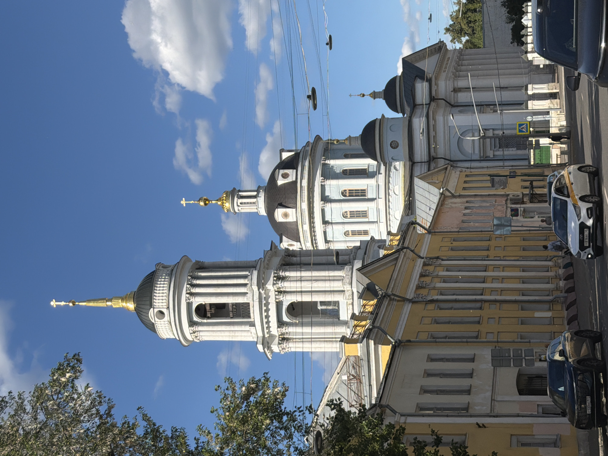 The image captures a street scene in Moscow, Russia, with a prominent church building dominating the frame. The church features multiple domes and architectural details characteristic of Russian Orthodox churches, including golden crosses atop its spires. The building's facade is painted a light blue and white, with a yellow-painted building adjacent to it. A tree with lush green foliage is visible in the foreground on the left.
Several vehicles are present on the street, including a white taxi with yellow markings, a dark blue car, and part of another vehicle on the far right. The sky is blue with scattered white clouds, suggesting a clear day. Electrical wires crisscross the sky above the street and the church. The overall scene appears to be during daylight hours, likely mid-morning or afternoon, given the bright lighting and clear sky. No people are clearly visible or engaged in any specific activity, with one figure appearing to be walking near a crosswalk sign. There is text visible on the license plates of the cars, but it is not legible.