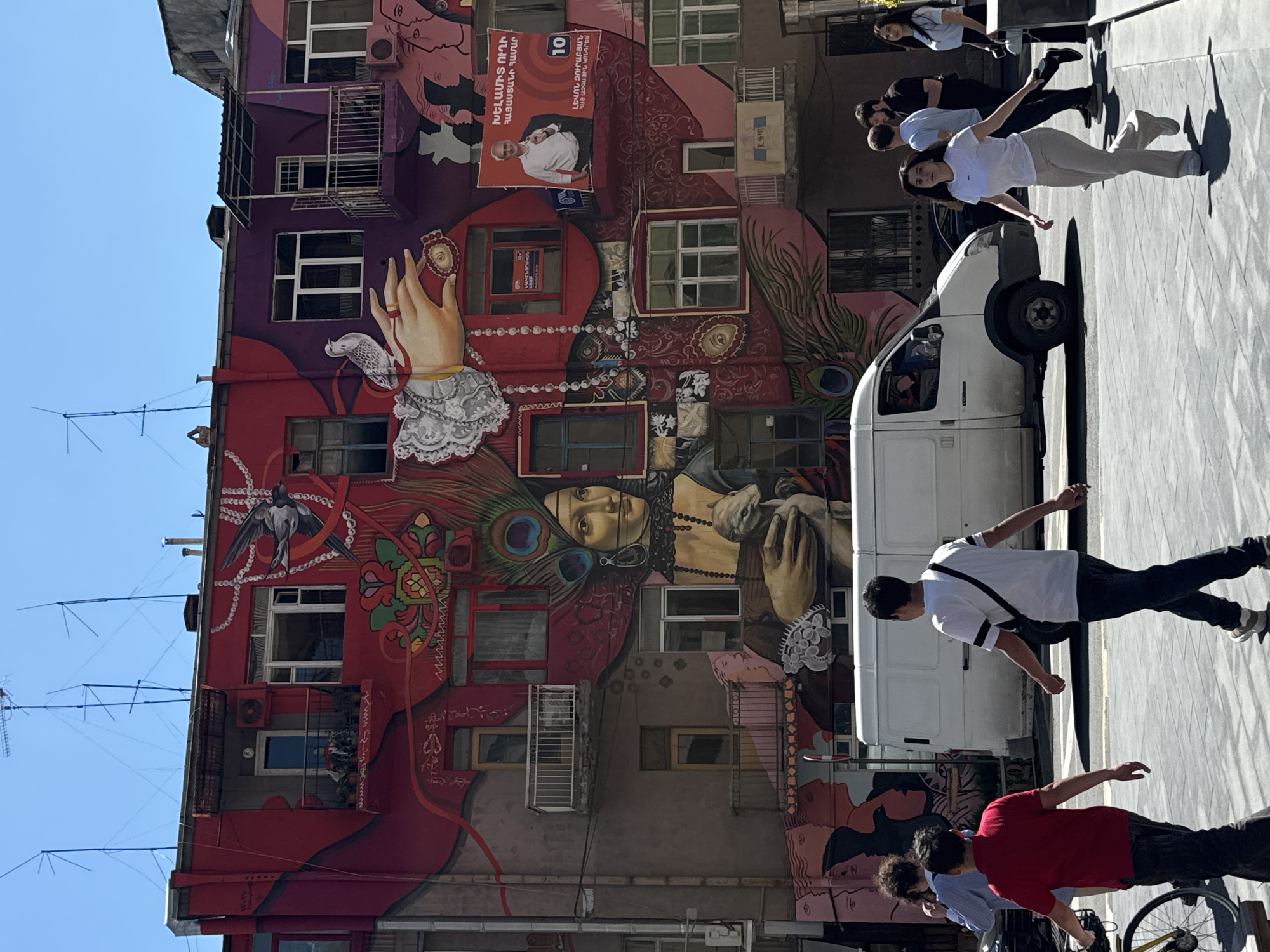 A photograph captures a Yerevan, Armenia, cityscape featuring a vibrant mural on the side of a building. The mural is rich in detail, portraying a woman with a serene expression adorned with peacock feathers and intricate patterns. A hand with lace cuffs and a ring on one finger holds an ornate object. Above, a political poster with a person in a white shirt is visible.
A white van is parked in the foreground, partially obscuring the mural. Several people are walking along the sidewalk; one woman wearing white stretches her arms outward and gazes upward. A man in a white shirt and dark pants walks towards the right, while another person in a red shirt is towards the bottom of the image, seemingly riding a bicycle.
The mural's colors range from deep reds to purples, enhancing the building's exterior. The sky is clear and blue, providing a contrasting backdrop to the colorful scene. Visible antenna systems are atop the building, and various windows provide a sense of the building's structural layout.