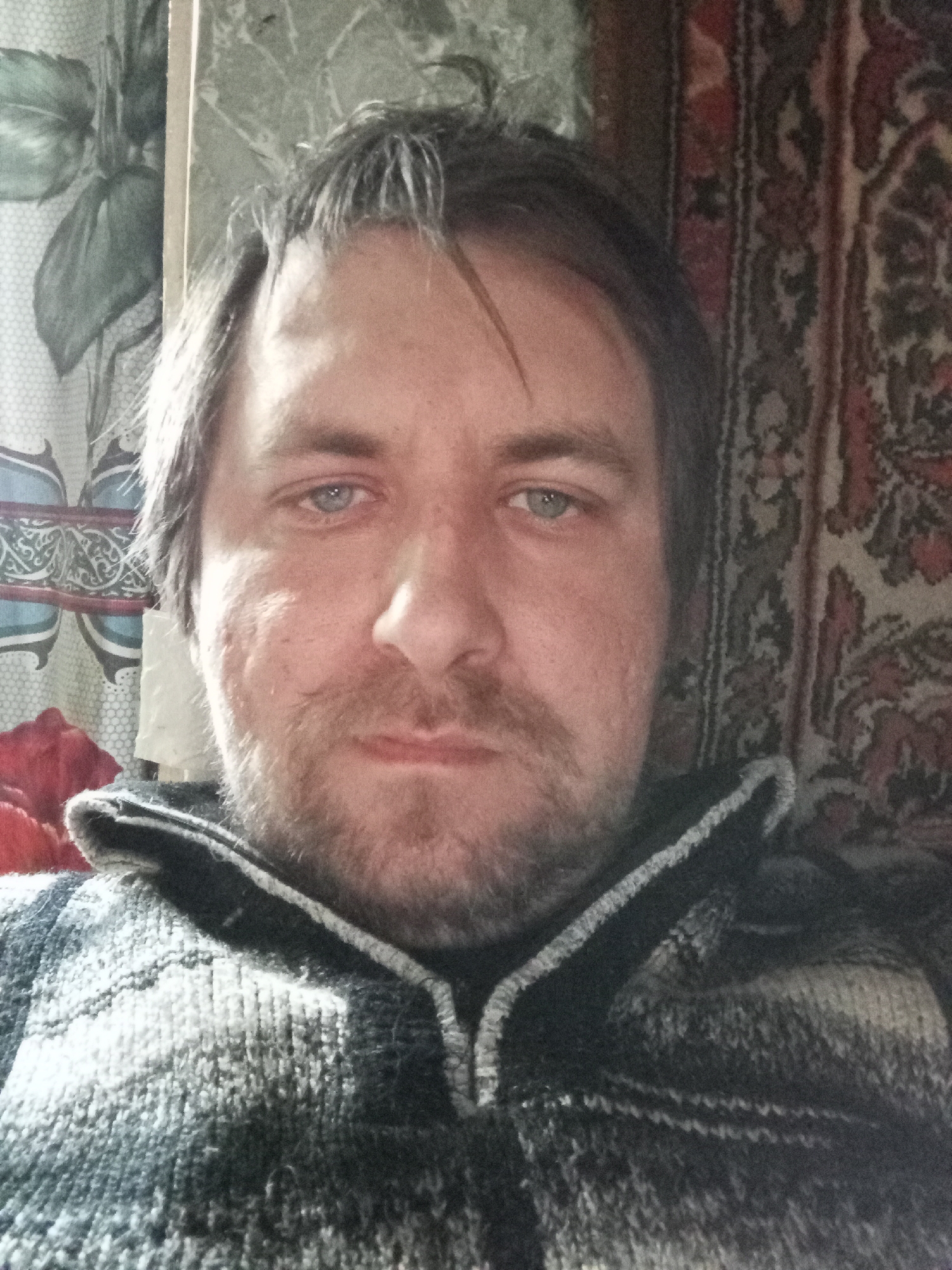 A close-up selfie of a middle-aged man with fair skin, light blue eyes, and a short beard, wearing a grey and black patterned sweater. The background includes a wall with floral patterned wallpaper on the left and a patterned rug on the right. There is no verifiable information to suggest that this photo was taken in the city of Parabel, Russia.