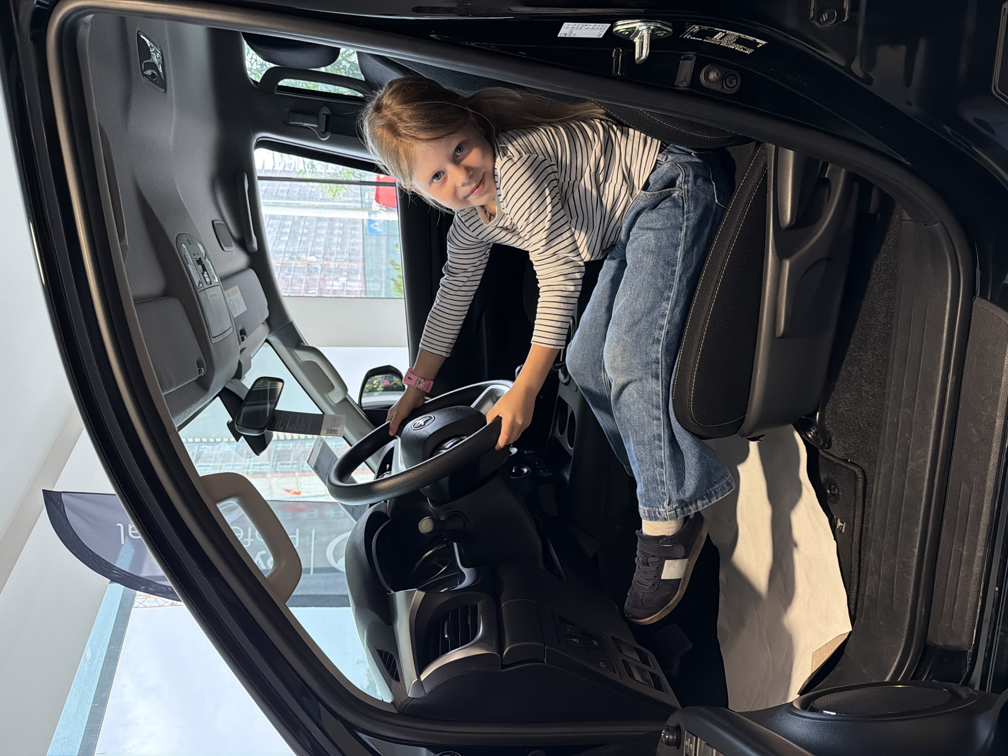 A young girl is positioned inside the cabin of a truck. She is fair-skinned with blonde hair and blue eyes, wearing a striped long-sleeved shirt, blue jeans, and dark blue shoes. She is holding the steering wheel and looking directly at the camera.
The truck's interior is black. A white protective sheet covers the vehicle's interior. The background shows a blurred storefront and cityscape.
The location is Podgorica, Montenegro.
