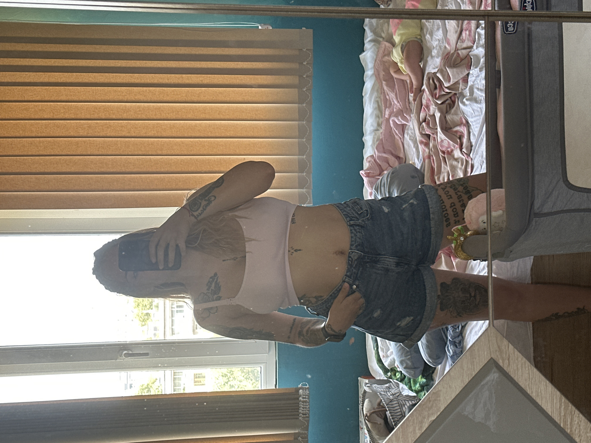 The image shows a woman in denim shorts and a pink crop top standing in front of a mirror in what appears to be a bedroom. She is taking a selfie with her phone. She has tattoos on her arms, torso, and legs. In the background, there is a window with blinds, a blue wall, and a bed with blankets and a baby lying on it. A baby crib is also visible in the foreground. The scene appears to be indoors, possibly a residential apartment. The city of Plovdiv, Bulgaria is not mentioned in the image.