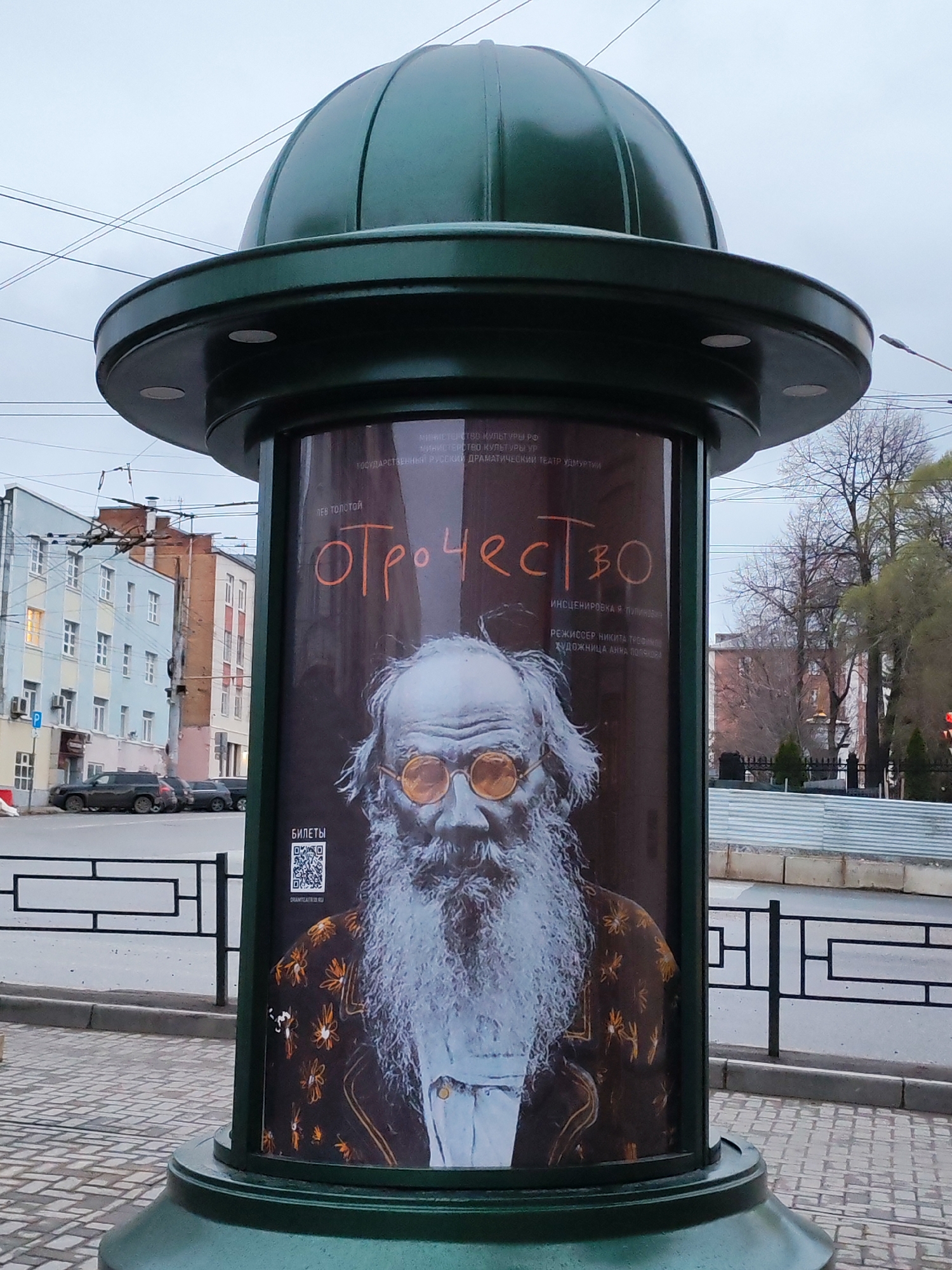 A cylindrical green advertising pillar stands on a cobblestone sidewalk next to a street in Izhevsk, Russia. The advertisement on the pillar is for a theater production of Leo Tolstoy's "Childhood". It features a grayscale portrait of a man with a long white beard and round yellow glasses. The text on the advertisement is in Russian. Behind the pillar, there is a street with cars, buildings, trees, and a metal fence. The sky is overcast.