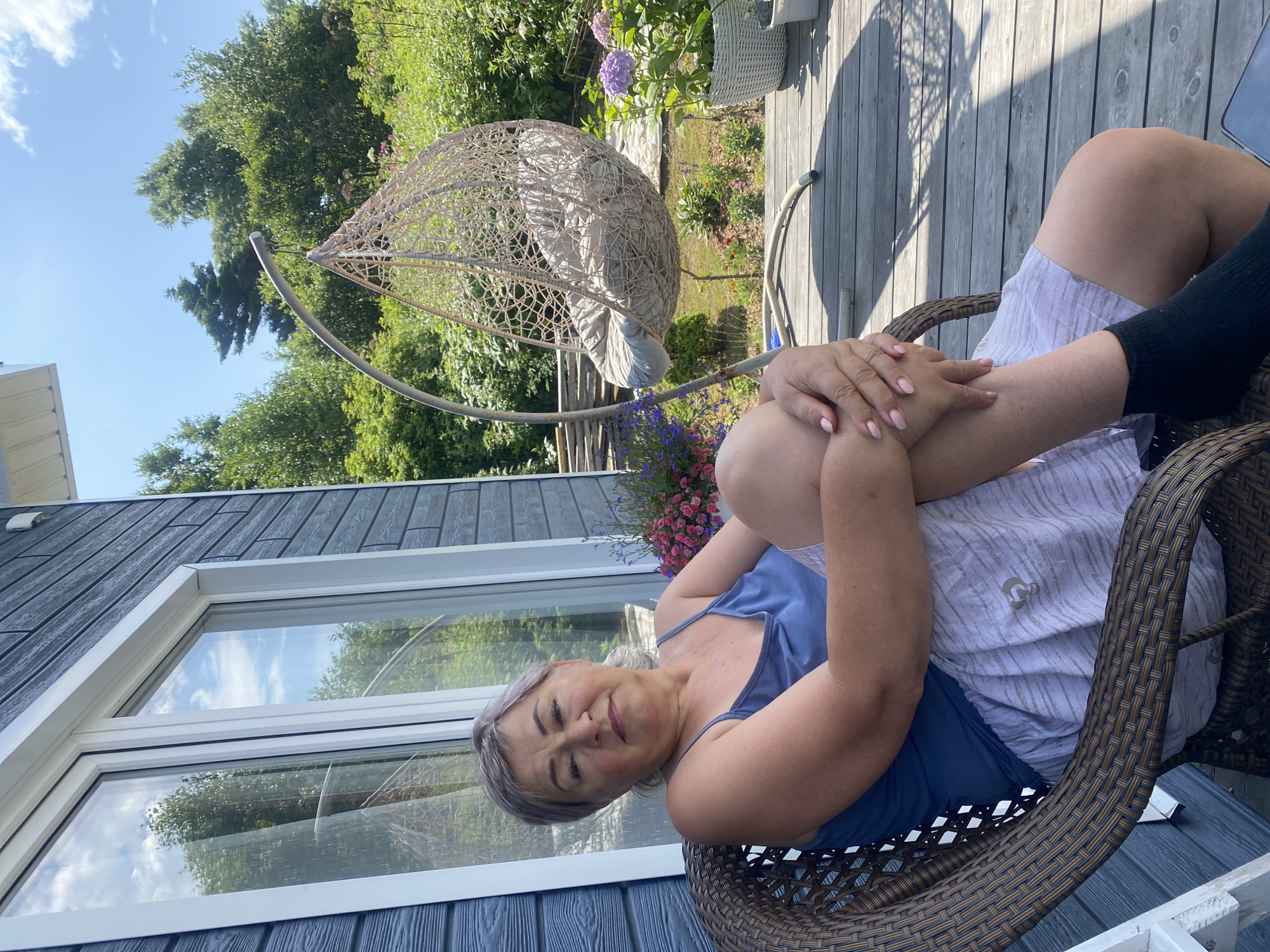 A middle-aged woman with short gray hair is sitting on a wicker chair on a wooden deck. She is wearing a blue tank top and light-colored shorts with a patterned design. Her arms are crossed and her legs are drawn up. Behind her, a hanging rattan chair is suspended from a metal stand. The deck overlooks a garden with various plants and flowers, including hydrangeas. A house with dark blue siding and a large window is visible on the left. The sky is clear and blue with some white clouds. The image appears to be taken in a residential area, likely in a rural setting given the lush greenery. The location of Tret’ya Pad’, Russia is not discernible from the image.