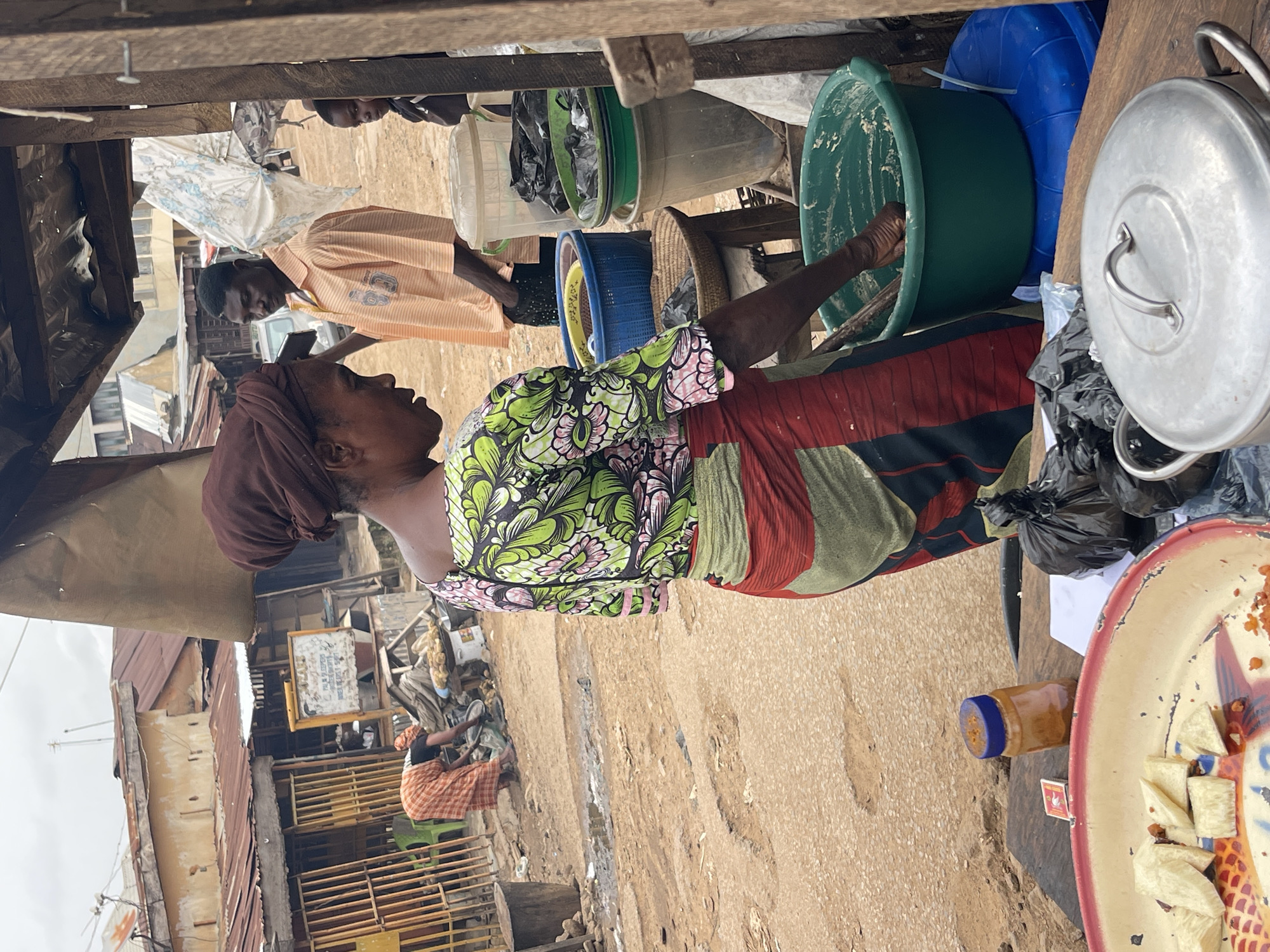 Here is a detailed description of the image:
**Location:** The image appears to be taken in a bustling, possibly low-income area of Chikaji, Nigeria. The setting is an outdoor market or street-side vending area.
**Environment:** The environment consists of a dirt or gravel ground and makeshift structures of wood and corrugated metal that serve as shops or dwellings.
**People:** The scene contains multiple individuals:
* **Vendor:** A woman wearing a head wrap, a colorful floral top, and a striped skirt is seen standing behind a table or display. She appears to be interacting with a food preparation process, possibly mixing ingredients in a green plastic bowl.
* **Customer:** A man in an orange patterned shirt appears to be a customer.
* **Other Individuals:** There are other people in the background; their actions or involvement are not clear.
**Objects:** The area is crowded with various objects related to commerce and daily life:
* **Food Items:** A tray of white cut food (possibly yams or plantains) and a jar of what could be peanut butter are visible.
* **Cooking and Storage:** Plastic tubs, bowls, and a metal pot with a lid indicate food preparation or storage.
* **Market Display:** The vendor's stand includes a variety of plastic containers and miscellaneous items.
**Actions and Interactions:**
* The vendor is actively engaged in food preparation or sale.
* The man seems to be looking at something being displayed.
* The background shows activity that is typical of a busy market area.
**Overall Impression:** The image shows the daily activities of market life in Chikaji, Nigeria. The scene suggests a dynamic, commercial environment where people are involved in buying, selling, and preparing food. The infrastructure is modest, reflecting the economic circumstances of the area.