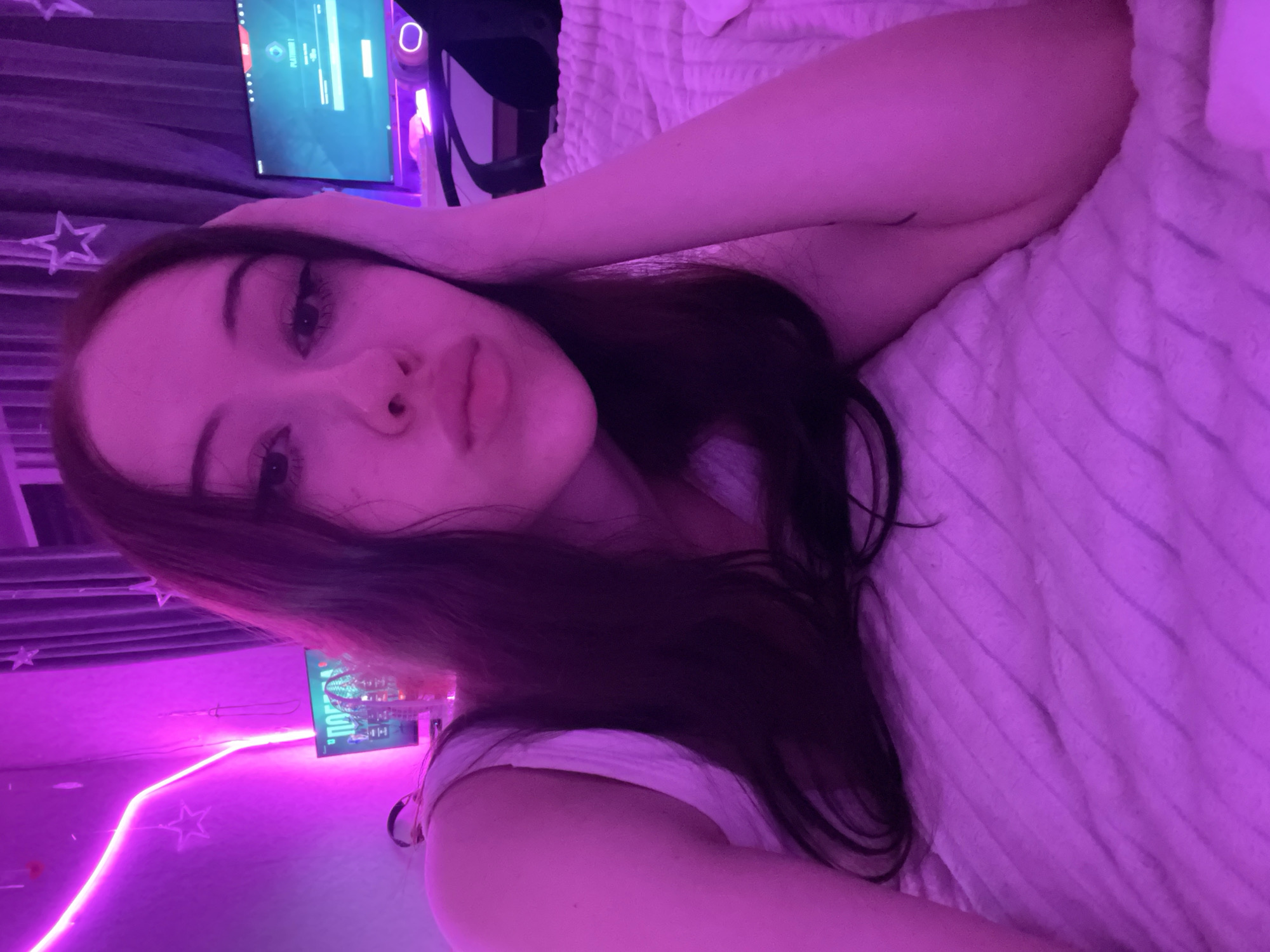 A young woman with long dark hair is lying on a bed with a textured pink comforter. She is looking at the camera with her head resting on her hand, which is propped up behind her head. Her eyes are open, and she has makeup on. She is wearing a white tank top.
In the background, there are two computer monitors. The monitor on the left displays a video game with colorful graphics. The monitor on the right displays a login screen with the text "PLATFORM 1". There is a pink neon light strip running along the wall on the left side of the image. The room is illuminated with pink light, creating a vibrant and atmospheric setting. Curtains with star patterns are visible in the upper right corner of the image. This scene appears to be in a bedroom, possibly a gaming room, located in Votkinsk, Russia.