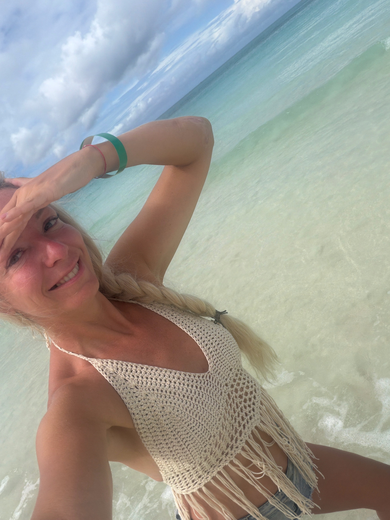 This is a selfie taken by a blonde woman at the beach in Malay, Philippines. She is smiling and wearing a cream-colored crocheted top with fringe and denim shorts. She has a green bracelet on her left wrist and her right arm is raised, shielding her eyes from the sun. The water is crystal clear and shallow, with gentle waves lapping at her legs. The sky is partly cloudy, suggesting it could be any time of day, though the lighting implies it's likely daytime. The overall mood of the image is relaxed and happy, conveying a sense of enjoyment of the tropical beach setting.