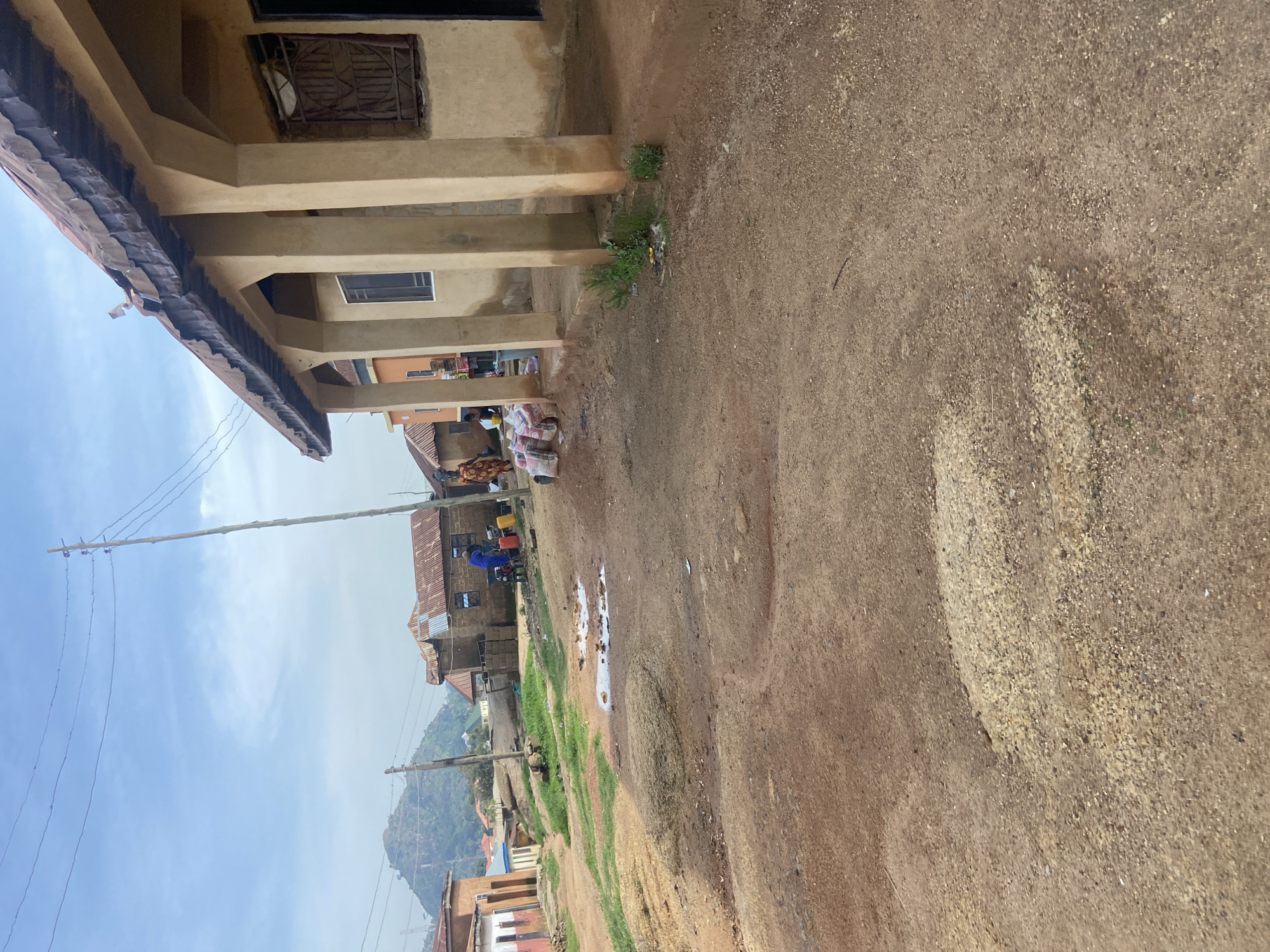 This image depicts a scene in the city of Jos, Nigeria, focusing on a dirt road in front of a building with a covered porch. The building features a weathered, corrugated metal roof and a tan-colored facade with several supporting pillars. There are windows visible on the building, one with a metal grate.
In the foreground, the road is unpaved and consists of dirt and gravel. Puddles of water are present on the road surface, suggesting recent rain or a water source. Several large sacks, possibly containing goods or building materials, are piled near the porch of the building.
Further down the road, other buildings with corrugated metal roofs are visible, characteristic of many structures in the region. In the background, a prominent rocky hill or mountain formation rises, a common geographical feature in and around Jos. Electrical poles and wires traverse the sky, indicating the presence of infrastructure.
There are a few people present in the scene. One individual, dressed in a blue garment, appears to be interacting with equipment near the building. Another person, in a patterned dress, stands to the right of the frame. A few other figures are present in the distance, adding to the sense of everyday life in this location. The overall impression is a snapshot of a typical residential or commercial area in Jos, showcasing its natural landscape and urban environment.