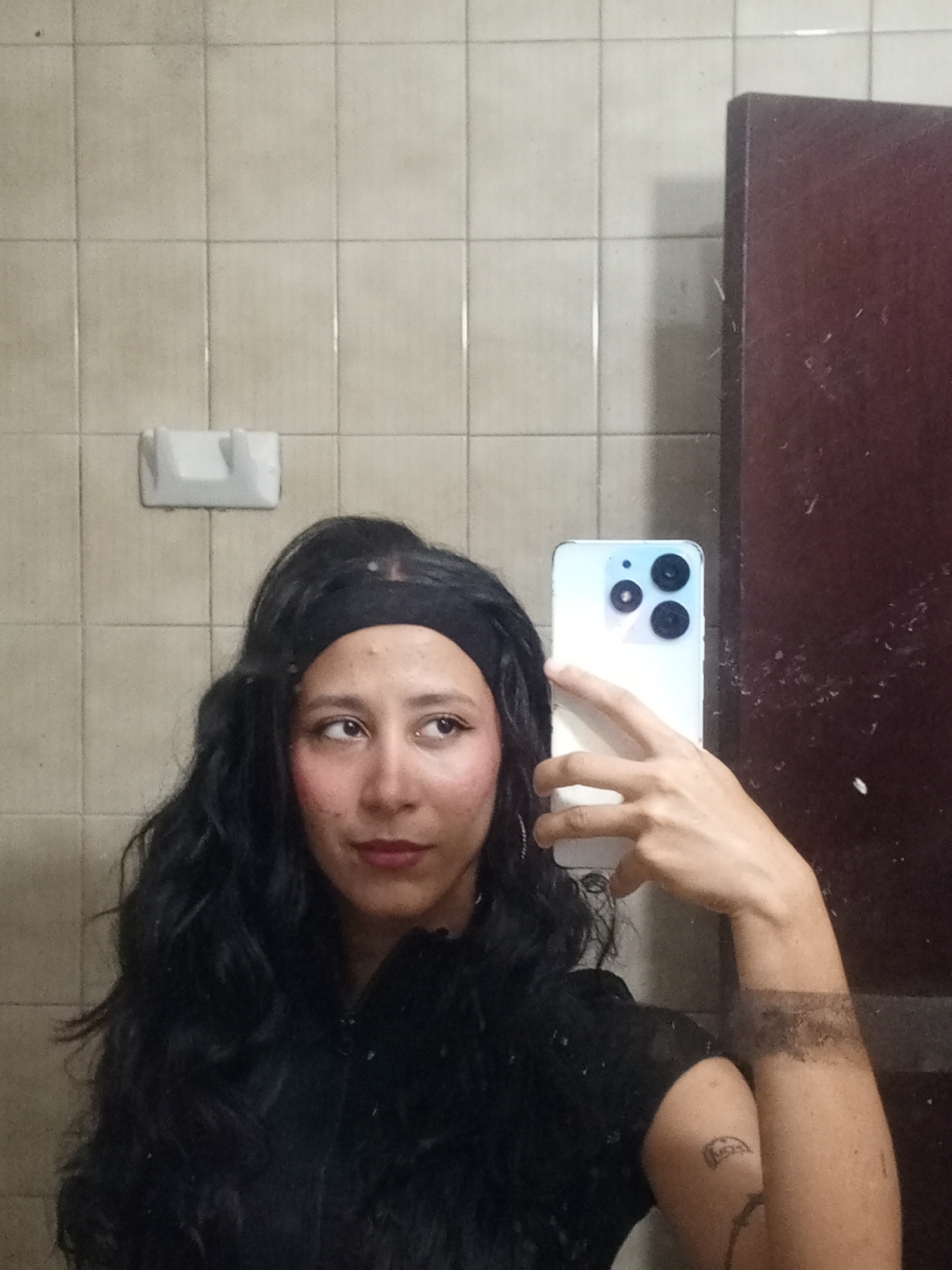 A woman takes a mirror selfie in what appears to be a tiled room, possibly a bathroom, in Porlamar, Venezuela. She has long, dark, wavy hair, styled with a black headband, and is wearing a black top. Her face is made up with pink blush and lipstick, and she gazes slightly to her right.
She holds a light-colored smartphone, featuring a prominent triple camera system, which partially covers her face. Visible on her left arm (viewer's right) are two tattoos: a dark band design around her forearm and a smaller, distinct tattoo on her upper arm.
The background consists of light-colored, square wall tiles with darker grout lines. A white, rectangular fixture, resembling a soap dish or holder, is mounted on the tiled wall behind her head. To the right of the frame, a dark brown, possibly wooden, surface is visible, which could be a door or a cabinet. The tiles and grout exhibit some signs of wear or age. No discernible text is visible in the image.