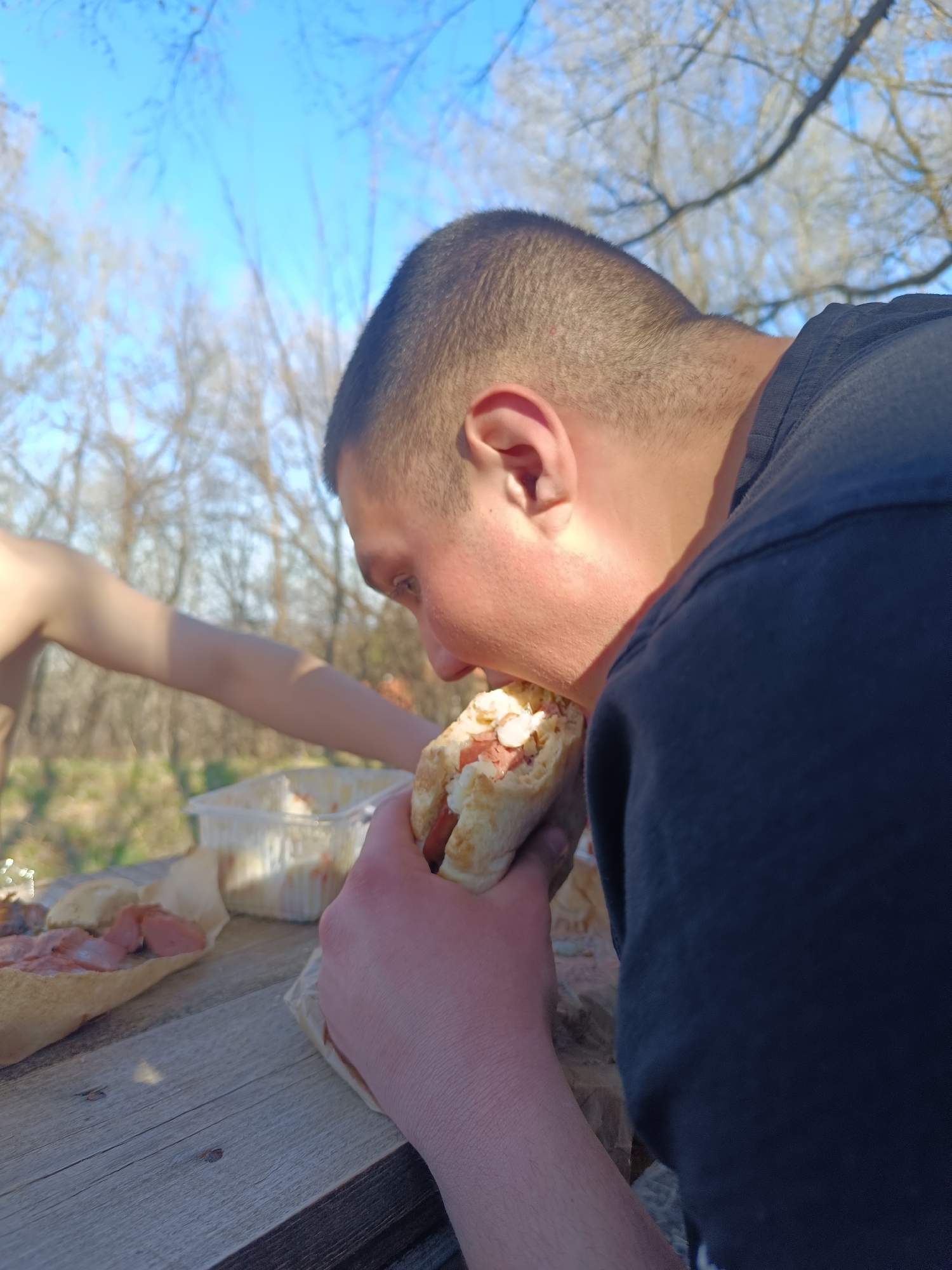 A man is eating a wrap outside on a sunny day. The man has short dark hair and is wearing a black shirt. He is holding the wrap in his left hand. There is a table with food on it in front of him, including a container of salad and a baguette with meat on it. Another person's arm is visible to the left. The sky is blue and there are bare trees in the background. The location is Unknown, Unknown.