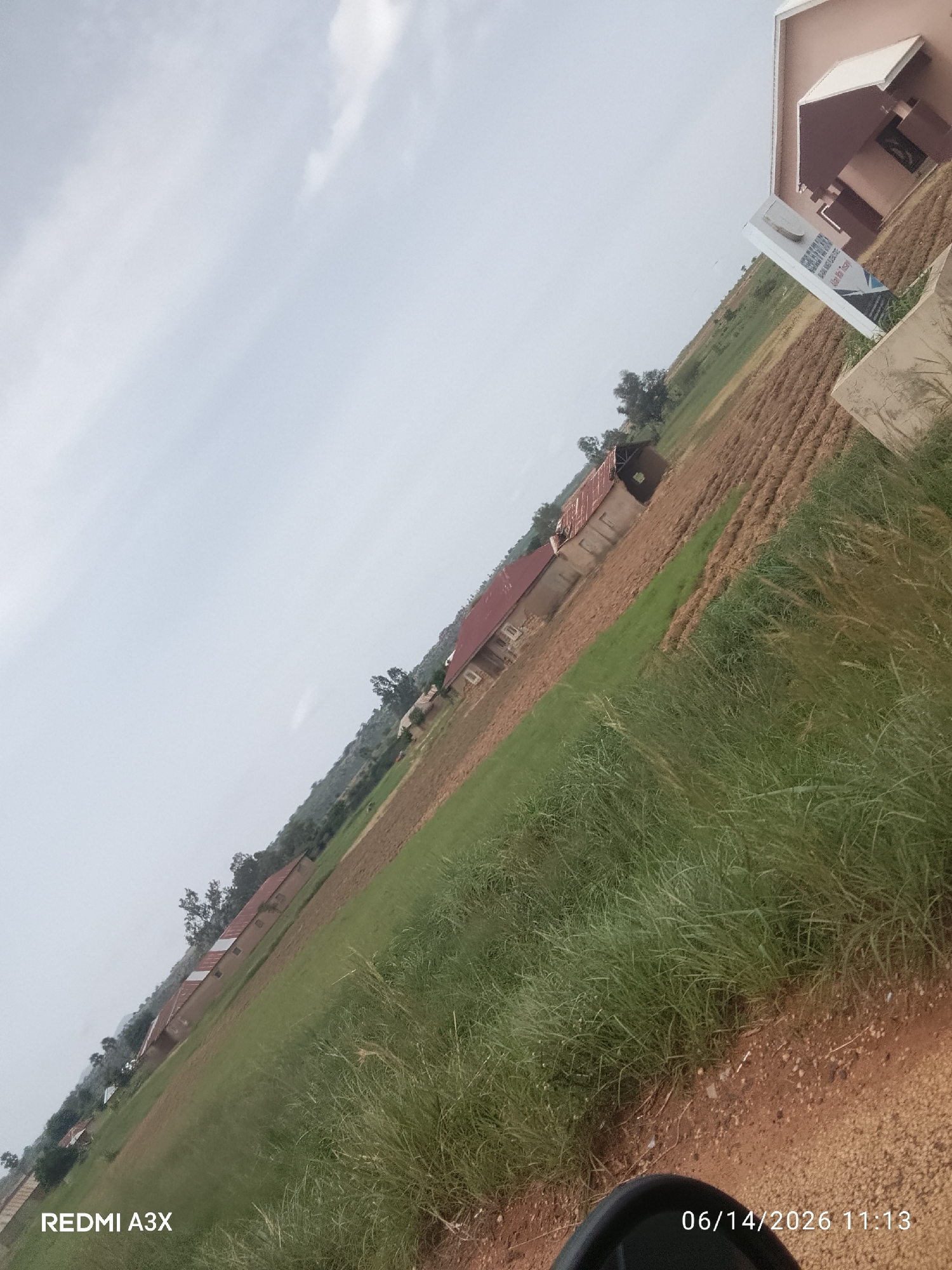 The image is an outdoor scene with a diagonal orientation, likely taken in Dorowa, Nigeria, as indicated by the timestamp which includes "06/14/2026". The foreground shows a patch of tall, green grass and a dirt road surface. In the mid-ground, there are several buildings with red roofs, appearing to be houses or small structures, situated amidst cultivated fields, some of which are brown earth and others are green. In the background, a larger, two-story building with a beige exterior and a white awning is visible. A white signpost with text and graphics is positioned in front of this larger building. The sky is overcast with some cloud formations. The overall environment appears rural or semi-rural, with agricultural land and residential or institutional buildings. There are no people visible in the frame. The timestamp "06/14/2026 11:13" and the text "REDMI A3X" are present in the bottom corners of the image.