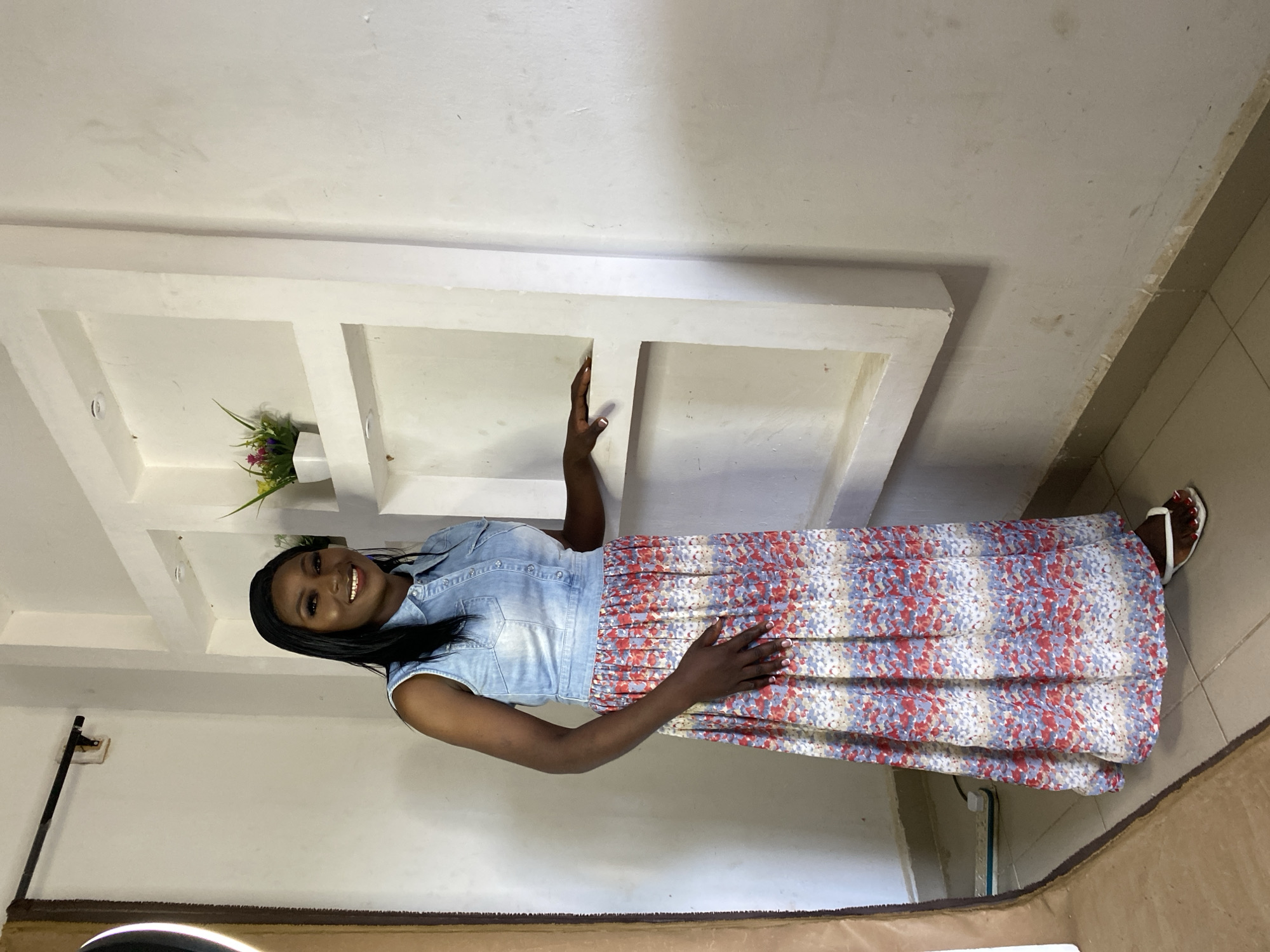 In an indoor studio setting, a Black woman stands in front of a white, multi-level shelf unit against a white wall. She is wearing a sleeveless light blue denim shirt over a long, flowing skirt with a floral pattern in shades of red, blue, and beige. Her right hand rests on the shelf, and her left hand is placed on her hip. She has dark hair pulled back and is smiling at the camera. White flip-flops are on her feet. The floor is tiled, and a tan-colored fabric hangs to the left of the frame. A power strip with multiple outlets is visible on the floor in the lower left corner. The lighting suggests a professional photo shoot.