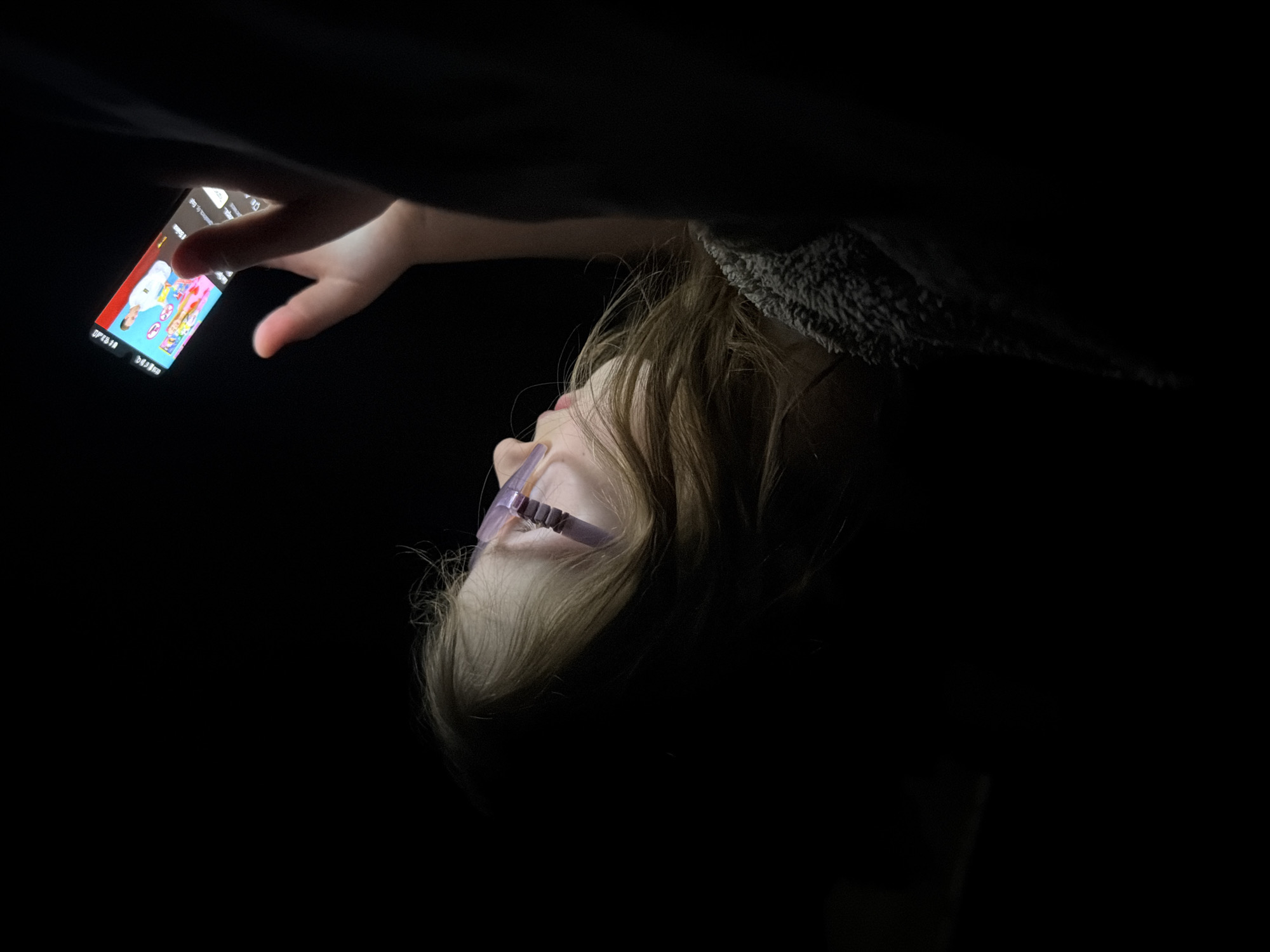 A child with blonde hair and glasses is lying down in a dark environment, illuminated by the light of a smartphone. The child is holding the smartphone with their right hand and appears to be interacting with the screen. The edge of a fuzzy grey blanket or jacket is visible near the child's head. The scene has a low-light quality, highlighting the child and the phone screen. There is no indication of location.
