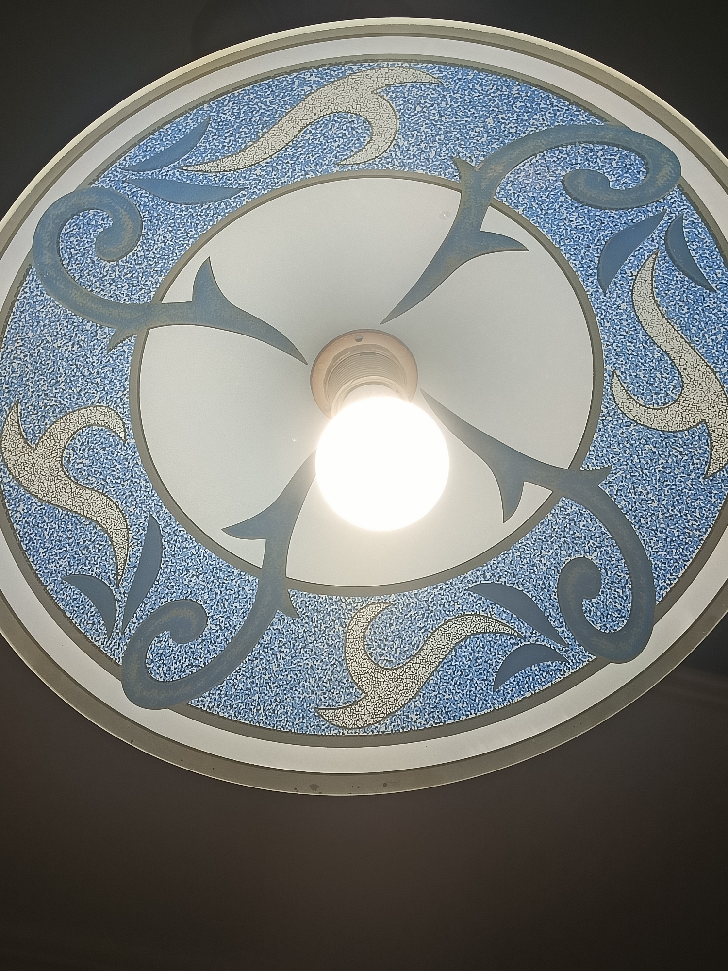 An upward angled shot shows a close-up of a circular, decorative light fixture with an illuminated lightbulb in the center. The fixture has a white central disc with a blue and white speckled border. Overlaid on this border are stylized, flowing designs in shades of blue and pale yellow. The lightbulb is emitting a bright white light, obscuring some of the detail of the fixture behind it. The background is a dark, nondescript color, suggesting the fixture is mounted on a ceiling or wall. This fixture is located in the lobby of the Varna Archaeological Museum.