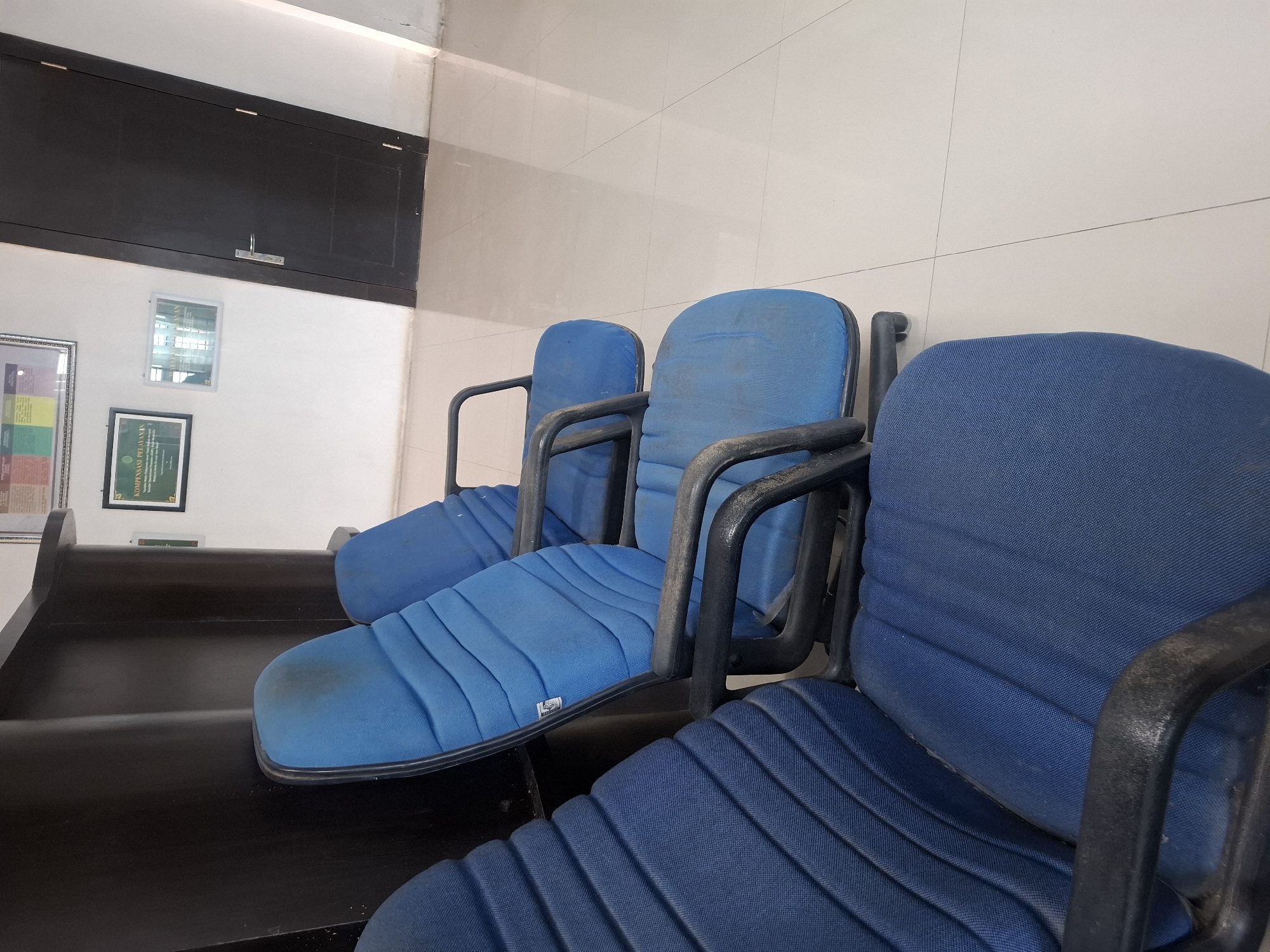 Three blue fabric chairs with black metal frames are positioned in a waiting area. The chairs are lined up in a row, with the closest chair occupying the foreground and the other two partially visible behind it. The chairs appear somewhat worn, with visible dust and some staining on the fabric. Behind the chairs, a white tiled wall runs horizontally across the background, marked by subtle grout lines. To the left, a dark wooden bench or seating area is visible. Mounted on the white wall above the bench are two framed documents. One is a certificate with text and a colored graphic, and another smaller frame contains an image. The overall environment suggests a functional waiting space, possibly in an office or public building. The location is identified as Meureudu, Indonesia, indicating an urban or semi-urban setting.
