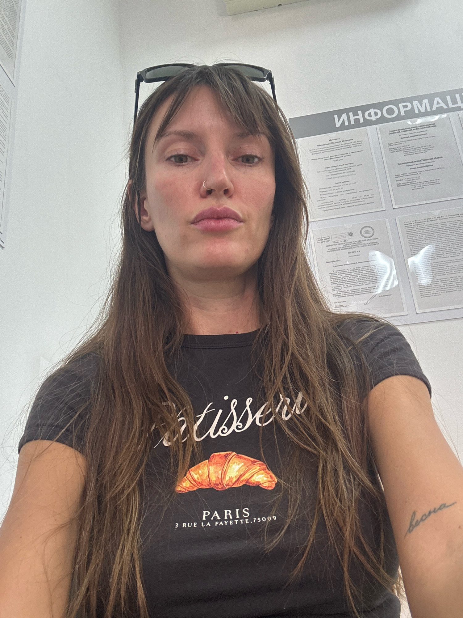 A young woman with long brown hair and bangs is shown from the chest up, taking a selfie. She is wearing sunglasses on top of her head and a dark t-shirt with a graphic of a croissant and the word "PARIS" printed on it. She has a nose ring and a tattoo on her right forearm that appears to say "весна". Behind her, there is a bulletin board with various documents and signs, one of which has the word "ИНФОРМАЦИЯ" visible at the top. The setting appears to be an indoor office or public service building. The information on the bulletin board, including official seals and Russian text, suggests a location within Russia. Specifically, the text on the bulletin board, mentioning a notary and addresses within the Rostov Oblast, allows for the inference that the location is in the city of Bataysk, Russia.