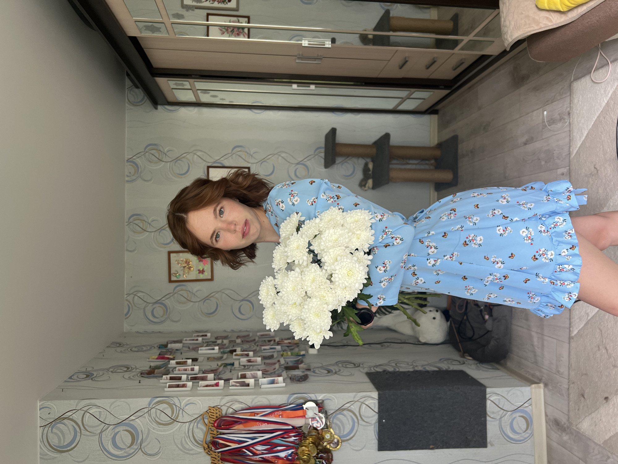 A young woman with shoulder-length auburn hair and blue eyes stands in what appears to be a domestic interior, possibly in Chaikovsky, Russia, given the context. She is wearing a light blue dress adorned with a floral and butterfly pattern. In her arms, she holds a large bouquet of white chrysanthemums. The background reveals a room with patterned wallpaper featuring grey and blue swirls. To the left, a wall displays numerous framed photographs and a rack of medals. A tall, dark grey cabinet or piece of furniture is visible, with a light-colored stuffed animal and a cat bed in front of it. To the right, a large wardrobe with mirrored doors stands. A cat tree with multiple levels is also present in the room. The floor is covered with light-colored wood-effect laminate and a patterned rug. The scene suggests a casual, personal setting.