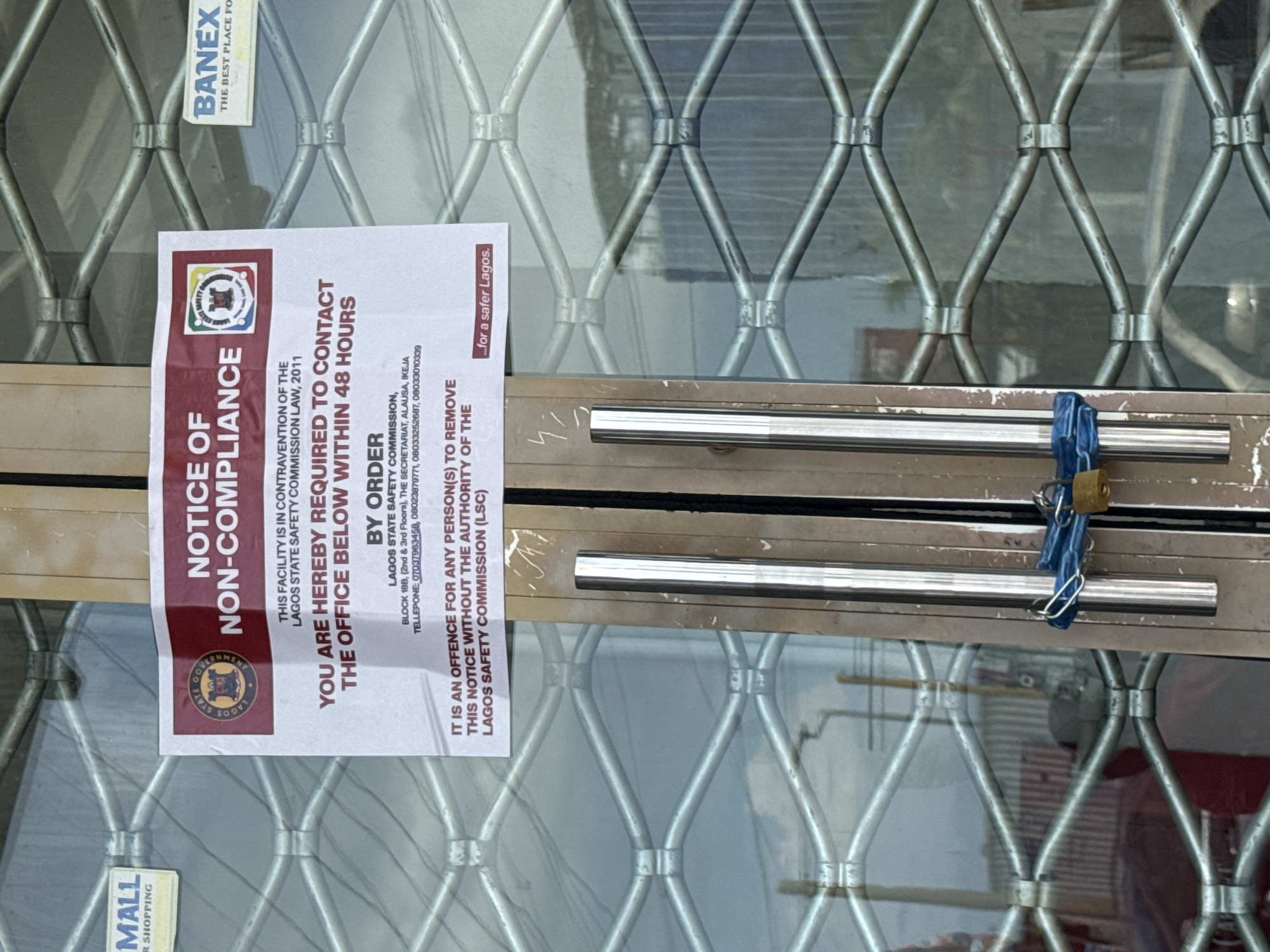 The image shows a locked storefront with a "NOTICE OF NON-COMPLIANCE" sign attached to the glass. The sign, in red and white, states that the facility is in contravention of the Lagos State Safety Commission Law, 2011, and requires contact with the office within 48 hours. It also warns that removing the notice without authority is an offense. The sign includes the Lagos State Government emblem and contact information for the Lagos State Safety Commission. The storefront is secured with a metal grate and a padlock, suggesting it is closed or has been sealed. The text "BANEX THE BEST PLACE FO" is visible on a sticker above the notice. A sign for "MALL R SHOPPING" is partially visible at the bottom left. The overall scene suggests a business that has been subject to enforcement action by a government agency. The lighting appears to be natural, indicating it's daytime.