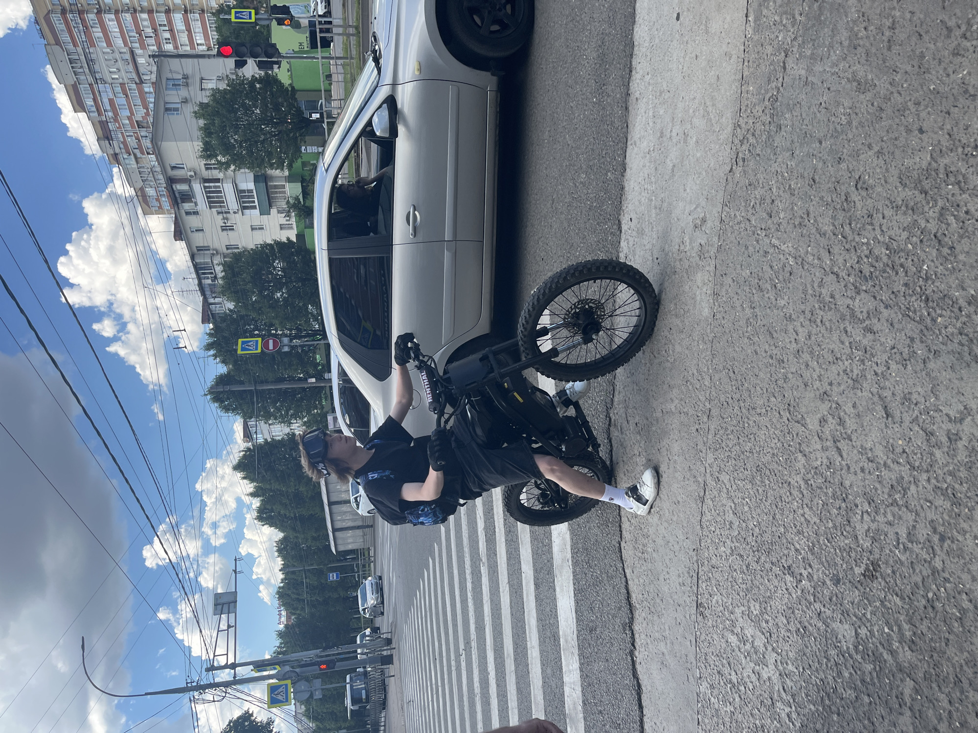 A person is sitting on a black electric motorcycle, which is positioned on a crosswalk. The person is wearing a black t-shirt, black shorts, white socks, and white and black sneakers. They are also wearing black gloves and a black headset or headphones. A small blue and black bag is slung across their chest. The motorcycle has a dark frame and spoked wheels.
The scene takes place on a street in Krasnodar, Russia. A light-colored car is parked next to the motorcycle, with its front passenger door open. The background shows a multi-story apartment building with many windows and balconies, as well as trees and a blue sky with white clouds. Traffic lights and power lines are also visible. The weather appears to be fair. The overall impression is of a casual urban scene on a bright day. The person on the motorcycle seems relaxed.