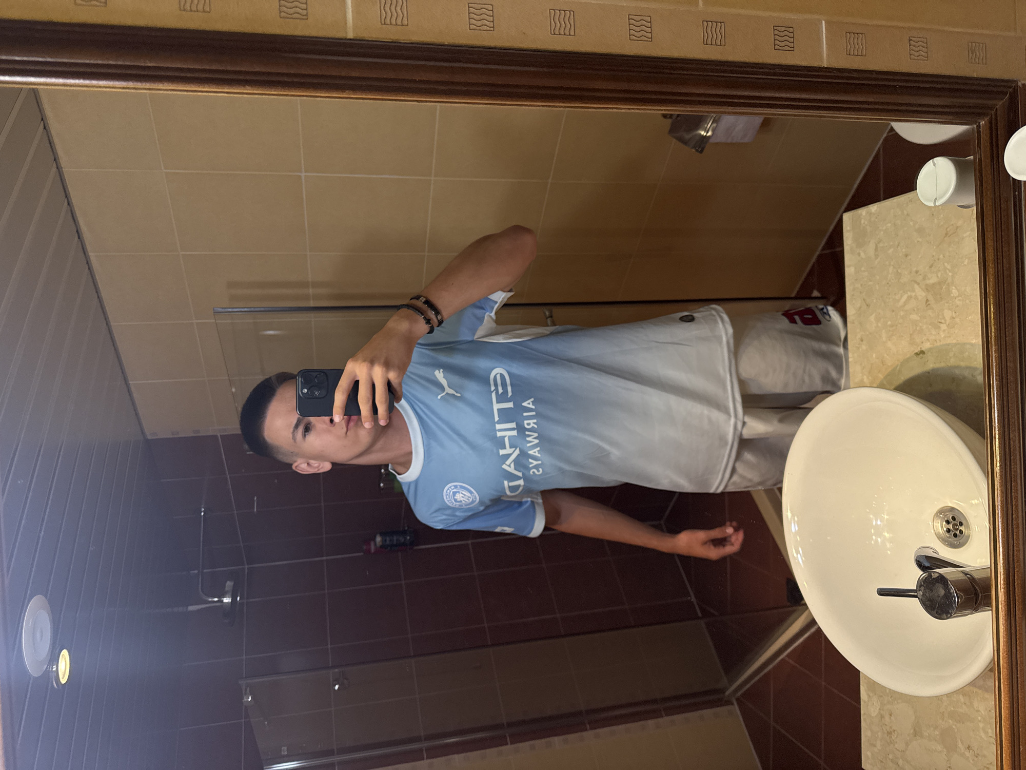 A young male is taking a selfie in a bathroom mirror. He is wearing a light blue and white gradient jersey and light-colored shorts. He has short, dark hair and is wearing two bracelets on his left wrist. The bathroom has beige tiled walls, a brown tiled shower enclosure, and a white sink with a chrome faucet. The mirror is framed in dark wood. The image does not contain any information that allows it to be placed in the city of Belogradchik, Bulgaria.