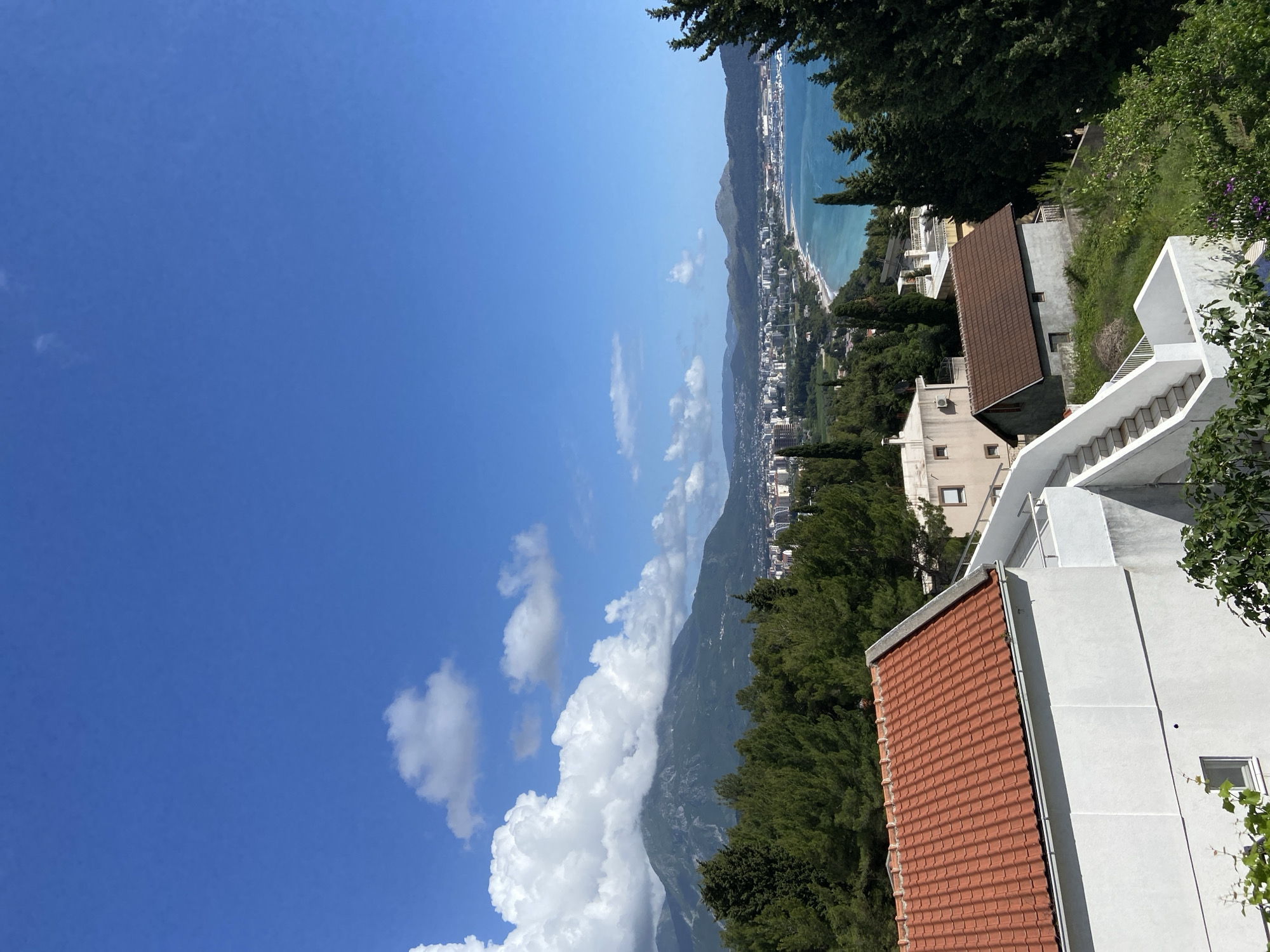 The image shows a view of Šušanj, Montenegro, from an elevated position. The scene is dominated by a clear blue sky with scattered cumulus clouds on the left. Below the clouds is a mountain range, partially obscured by trees. In the background, the Adriatic coastline is visible, with a strip of beach and a city stretching along the shore. Buildings with red-tiled roofs and white exteriors are positioned in the foreground and right side of the image, partly obscured by trees. A staircase is visible next to one of the buildings.