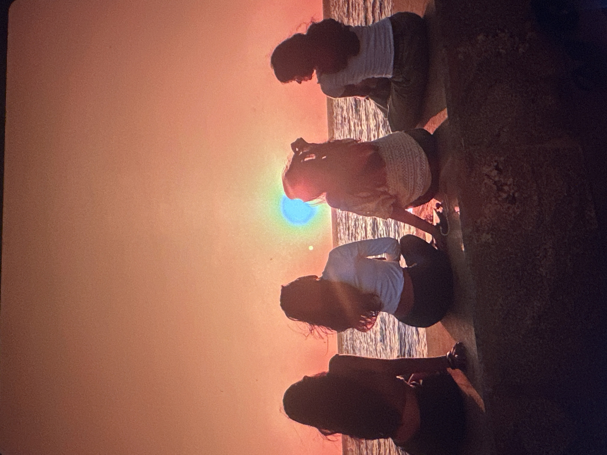 Four individuals are seated on a ledge, facing a body of water and a sunset. The sky is a gradient of orange and pink. The individuals are silhouetted against the bright sky, making it difficult to discern specific features, but they appear to be young women. The water is calm, with ripples visible. The ledge they are sitting on is made of concrete or stone. The background appears to be a natural landscape, likely a coastline. There is no visible indication of Tallinn, Estonia in the image.