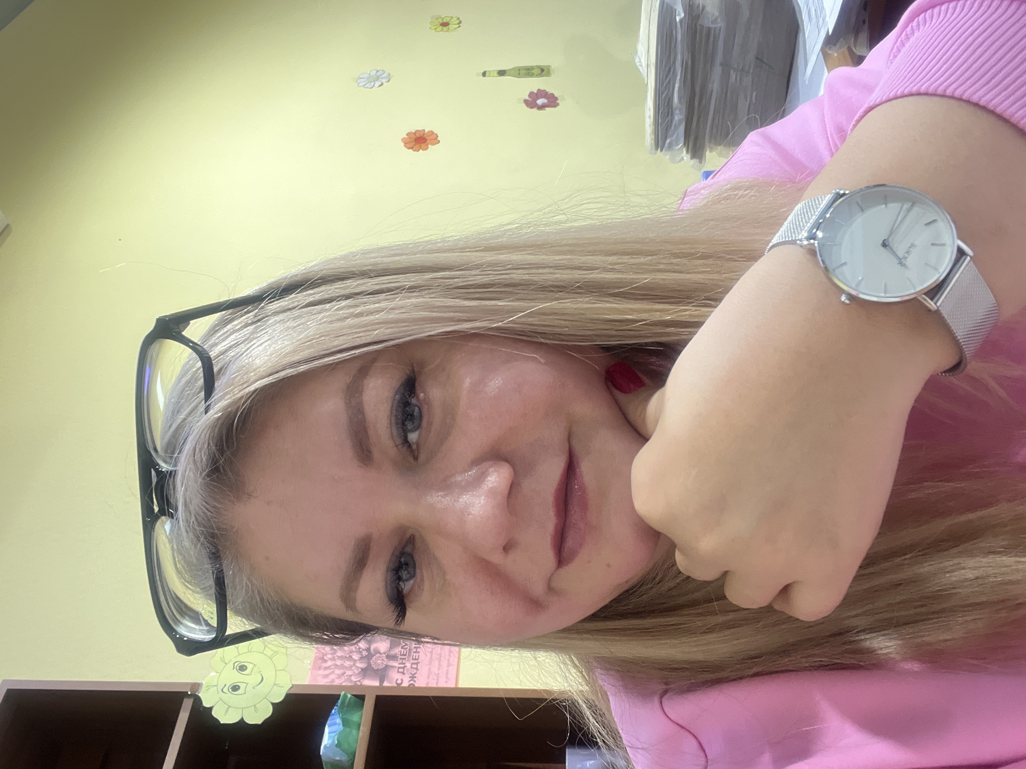 A fair-skinned woman with blonde hair is posing for a selfie. She is wearing glasses and a silver watch with a white face. She is wearing a pink sweater. Behind her is a yellow wall with several floral stickers and a green bottle sticker. A bookshelf with various items on it is visible in the lower left corner. The woman is holding her fist close to her face. There is no information in the image to determine the location as Izhevsk, Russia.
