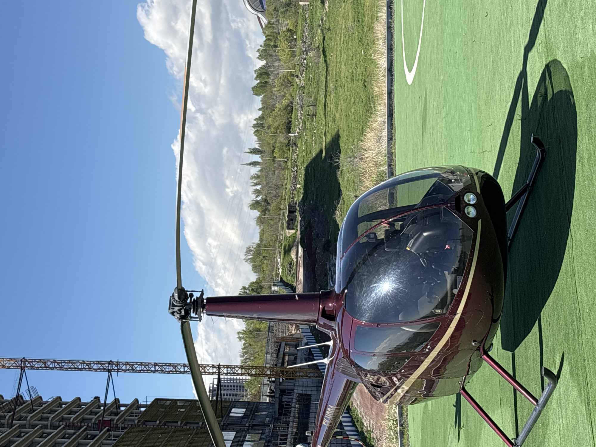 A maroon and beige Robinson helicopter is parked on a green helipad. The helicopter's windshield is cracked. A construction site with a crane and an unfinished building is in the background. In the distance, there are trees and light clouds in a blue sky. The scene appears to be in Yerevan, Armenia, based on the architecture and landscape.