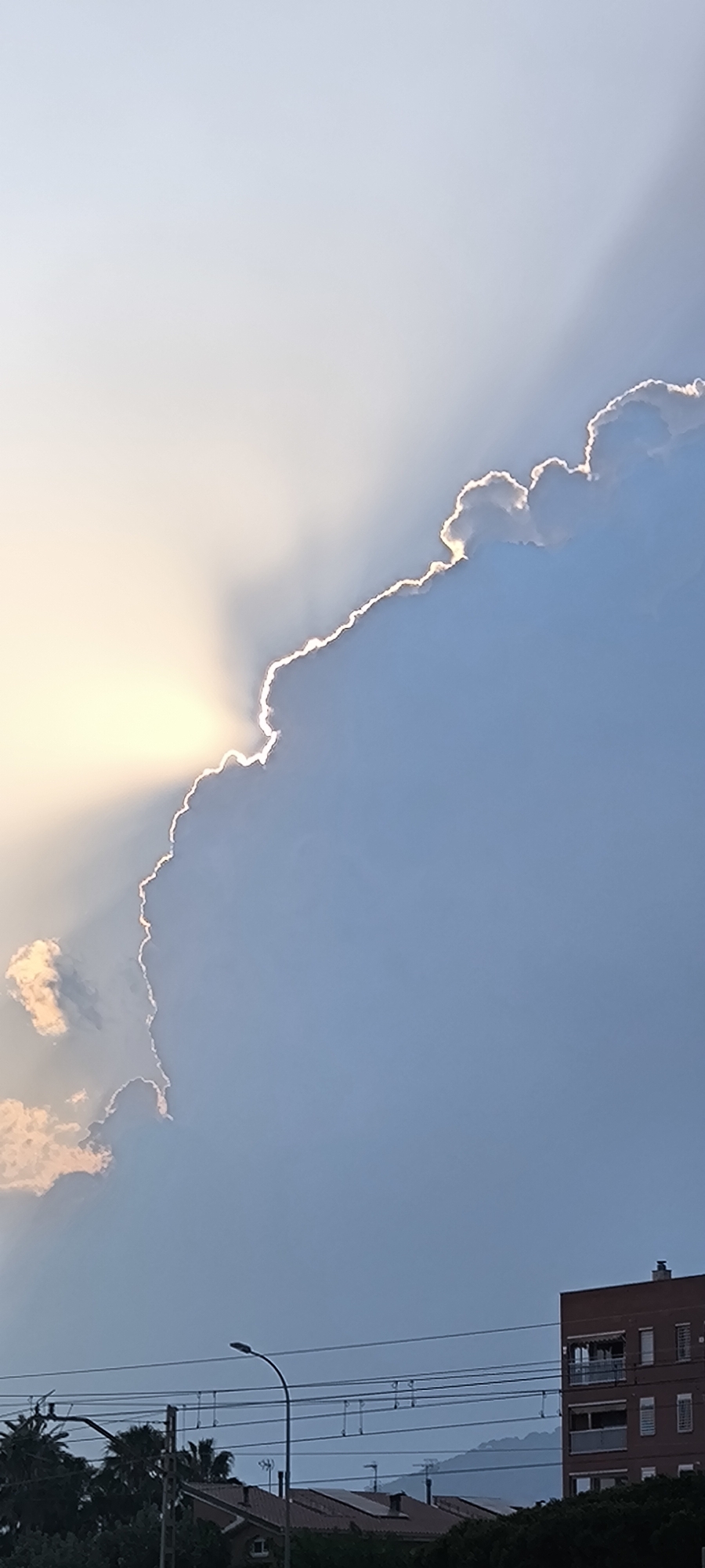 The image captures a dramatic sky over the city of Premià de Mar, Spain, with the sun's rays piercing through the edge of a large cloud, creating a bright, glowing halo effect. The sky transitions from a pale yellow near the sun to a soft blue. In the lower portion of the image, silhouettes of palm trees and building rooftops are visible, along with utility poles and power lines, suggesting an urban or semi-urban setting. A multi-story brick building with balconies is prominent on the right side. The overall atmosphere is serene and majestic, highlighting the natural beauty of the sky.
