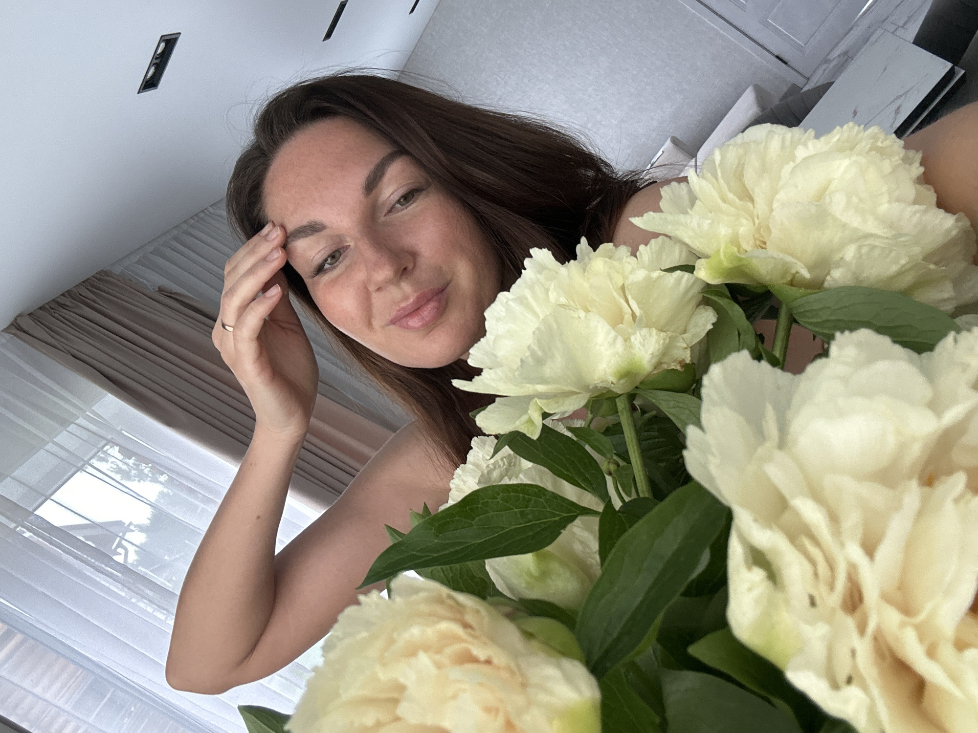 A woman with brown hair poses with a bouquet of light yellow flowers. She touches her forehead with her right hand. The background shows a window with white curtains and part of a room with a light-colored ceiling and furniture. The city is not discernible from this image.