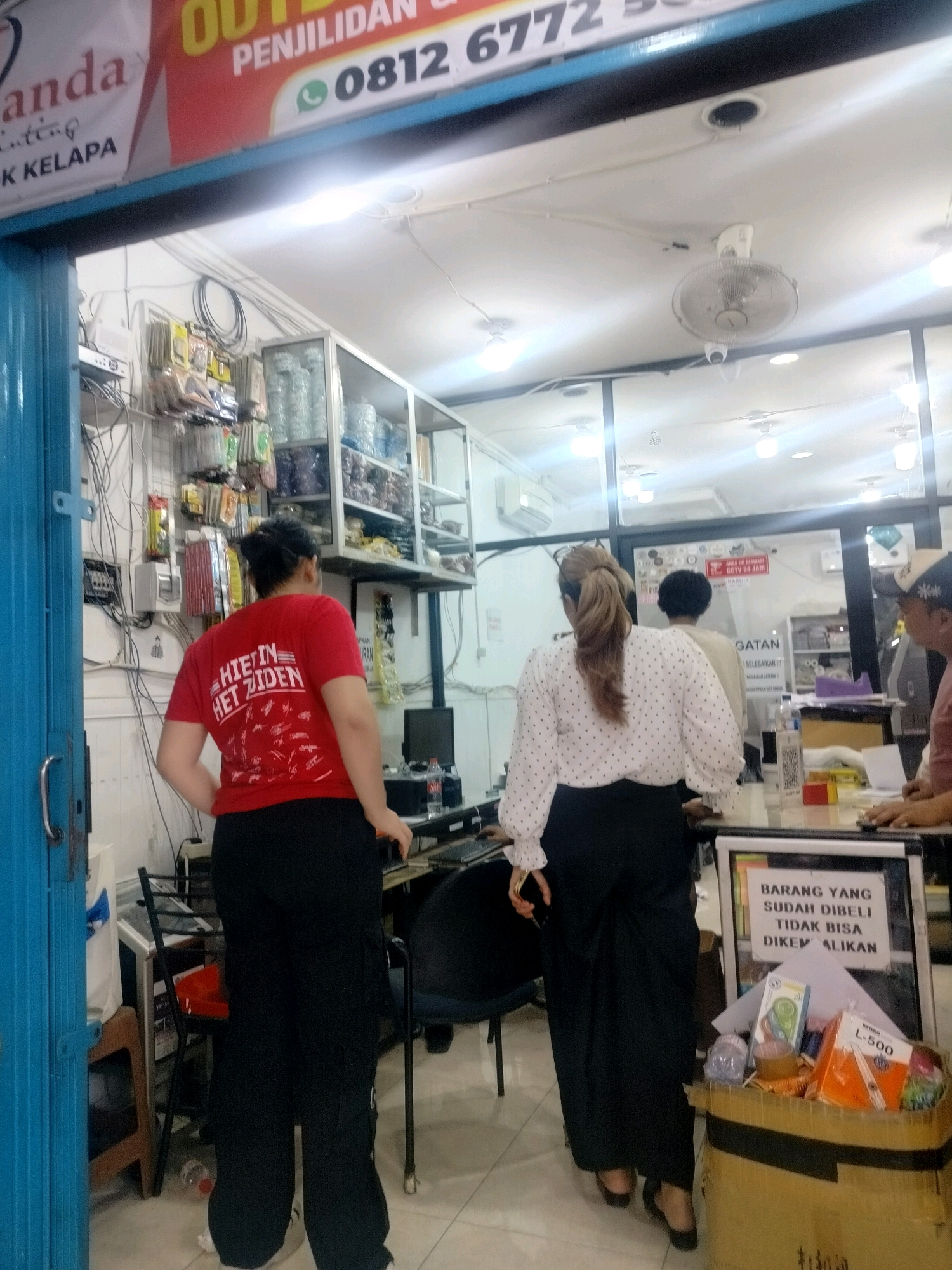 The image captures a view inside a store in East Jakarta, Indonesia, looking towards the interior. In the foreground, a person wearing a red t-shirt and black pants stands with their back to the camera. They appear to be looking at shelves stocked with various items. Further inside the store, another person in a white polka-dot shirt and black pants is also facing away from the viewer. A white board with text is visible near this person, and a cardboard box filled with items is on the floor. The store's interior features shelves, electrical wiring, and lighting fixtures. A ceiling fan is also visible. The overall impression is that of a retail space with merchandise and customers.