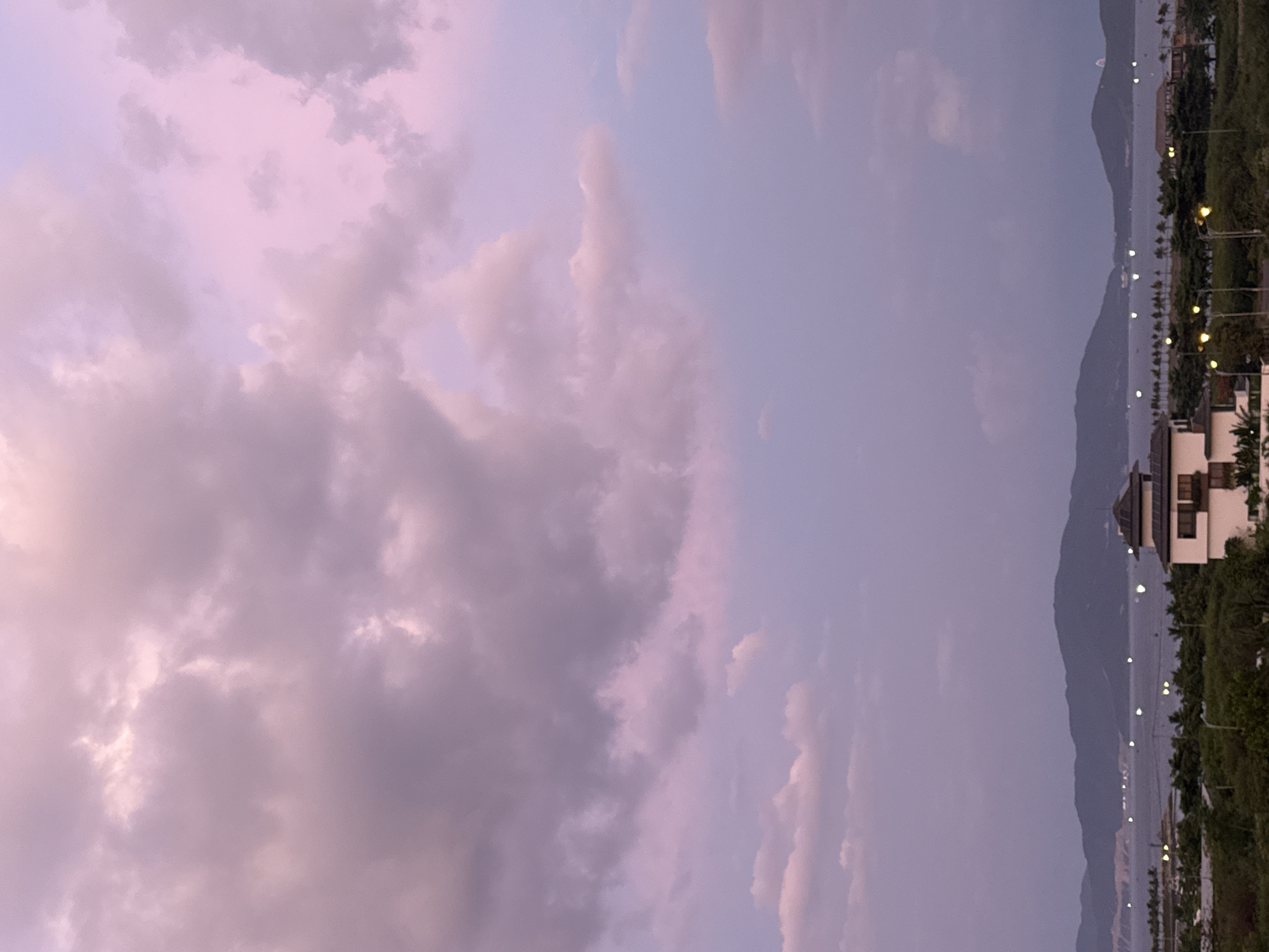 This image depicts a serene coastal landscape, likely taken in Nha Trang, Vietnam, during either sunrise or sunset. The sky is a prominent feature, filled with a dramatic and ethereal display of clouds. These clouds are tinged with soft hues of pink and purple, suggesting the warm glow of the rising or setting sun.
The scene transitions from the expansive sky to the horizon, where a mountain range forms a dusky silhouette against the lighter sky. Below the mountains, a body of water, possibly the sea, is visible, reflecting the ambient light. Along the coastline, there are hints of human habitation and infrastructure. Buildings, some with distinct architectural features, are nestled amongst lush green vegetation. The presence of streetlights, illuminated with a warm, yellow glow, suggests the twilight or early evening hours, with activity beginning to stir or wind down.
There are no visible people or specific objects that draw immediate focus, placing the emphasis on the natural beauty of the sky and landscape. The overall mood is peaceful and tranquil, evoking a sense of calm and natural wonder. The location cues – the mountains, the coastline, and the tropical vegetation – strongly indicate a seaside resort town. The weather appears calm, with the clouds suggesting fair conditions. No text is visible in the image.