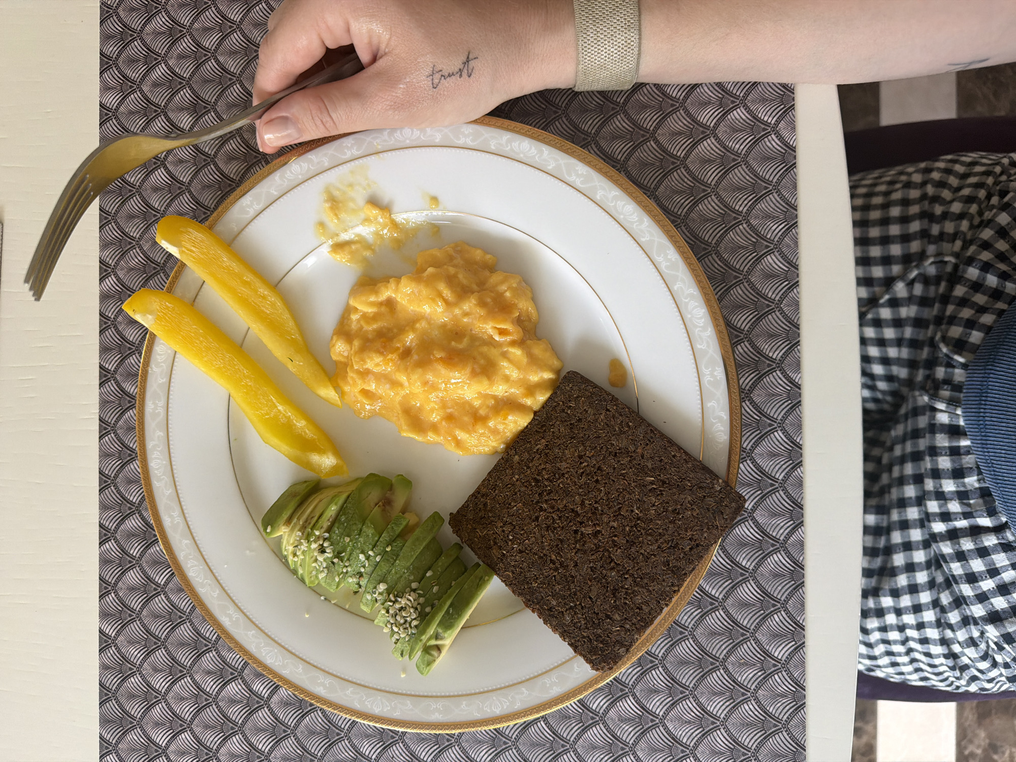 A plate of food is positioned on a table in what appears to be an indoor setting in Новоглаголево, Russia. The plate, which has a white surface with a decorative gold trim, contains scrambled eggs, sliced avocado with sesame seeds, yellow bell pepper strips, and a slice of dark bread. A hand holding a fork is visible. The wrist in the picture has a tattoo that reads 'trust' and is wearing a beige band. The table is covered with a patterned placemat. Part of a person seated at the table is visible, wearing a checkered shirt.