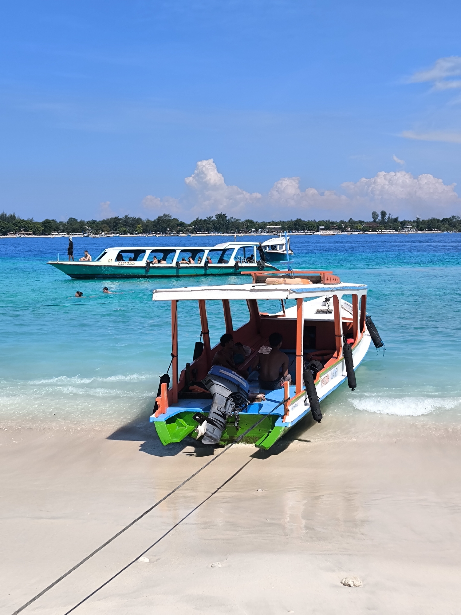 The image captures a vibrant coastal scene in an unknown city, Indonesia, showcasing two boats on a beach. In the foreground, a colorful boat rests partially on the sandy beach, its hull adorned with white, orange, green, and blue paint. Two individuals are seated inside the boat; their backs face the camera. An outboard motor is mounted at the stern. A long tow rope extends from the front of the boat onto the beach. In the background, a larger passenger boat floats in the turquoise water, carrying several people who are visible along the sides. Two individuals are swimming near the boat. The beach is white, and the water is clear and blue-green. In the distance, a strip of land is visible, covered with trees. The sky above is blue with scattered white clouds.