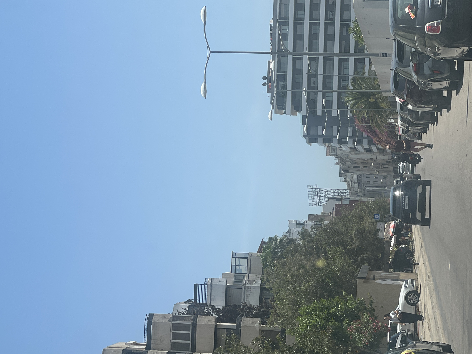 The image depicts a sunny urban street scene in Casablanca, Morocco. A clear blue sky dominates the upper portion. The asphalt road extends into the distance, with parked cars lining both sides. On the left, older multi-story residential buildings with tiered balconies and varying architectural details are partially obscured by mature green trees. On the right, a line of more modern, multi-story buildings with uniform grey facades and glass balconies stands behind a row of parked cars and prominent streetlights featuring double curved arms.
A black SUV, possibly a Range Rover, with a Moroccan license plate displaying "77981 | د | 7" is positioned centrally on the road, facing away from the viewer. A man wearing a dark red shirt and light shorts walks barefoot or in light footwear on the right side of the road, between the parked cars and the visible traffic lane, moving towards the distant end of the street. On the left sidewalk, a woman in a white top and dark pants stands near a parked white Volkswagen car. Other vehicles, including a motorcycle, are visible further down the street. A small, single-story structure or kiosk is present on the left sidewalk, and a large, skeletal billboard structure is visible in the distant background on the left side of the street. Pink flowering bushes and palm trees add greenery to the right side of the road.