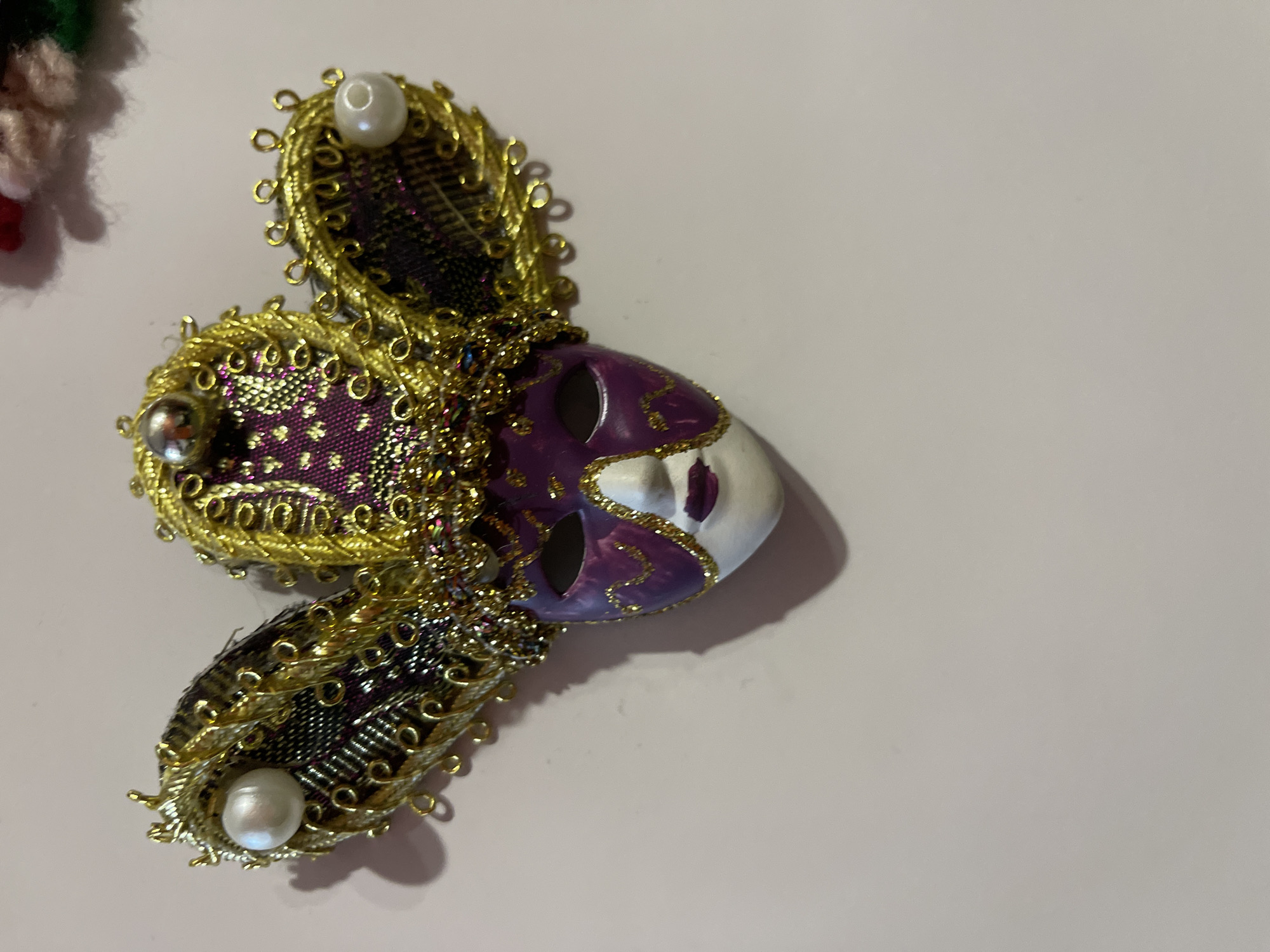 The image shows a decorative Venetian mask on a light pink surface. The mask is primarily purple and white, with intricate gold detailing and embellishments. It has a classic Venetian design with an elongated nose and eye openings. Three wing-like structures adorned with gold trim and pearls surround the central mask. These structures are made of a patterned dark fabric. The mask casts a shadow on the surface, indicating a light source. The location, Elhovo, Bulgaria, is not discernible from the image alone.