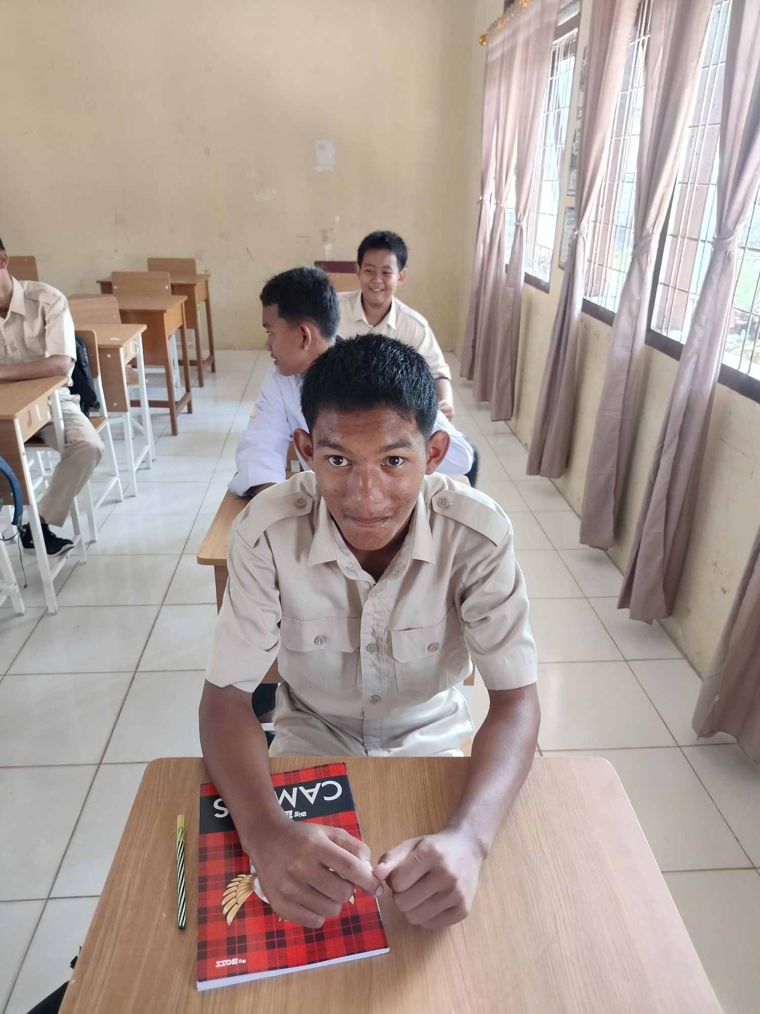In a classroom in Purwodadi, Indonesia, a group of male students in light brown uniforms are seated at wooden desks. The focus is on a student in the foreground, looking directly at the camera with a slight smile. He is wearing a short-sleeved, collared shirt with two epaulets on the shoulders. On his desk, there is a red and black checkered notebook with the word "CAMPUS" visible on the cover, and a pen with a striped design. Behind him, other students are seated in rows, with desks and chairs neatly arranged. The room has light-colored walls and large windows covered by beige curtains, allowing natural light to filter in. The tiled floor is a light cream color. The overall impression is of a typical school environment during a lesson or break.