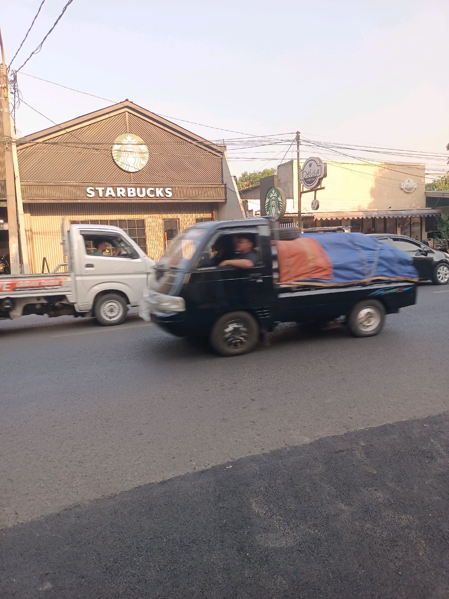 This is a photograph taken from a street-level perspective in Purwakarta, Indonesia. In the background, a Starbucks establishment is visible, characterized by its distinctive green siren logo and "STARBUCKS" lettering. Adjacent to the Starbucks is another building with signage that reads "DELISA delicious". The foreground features a street with two vehicles in motion. A white pickup truck is traveling from left to right, and a black pickup truck, loaded with blue and orange tarpaulin-covered cargo, is also moving in the same direction. The black pickup truck appears to be slightly ahead of the white one. The lighting suggests it is daytime, possibly late afternoon, with the sun casting long shadows. The sky is clear and light blue. The road surface is asphalt, with a clear division between newer and older paving visible in the lower portion of the image.