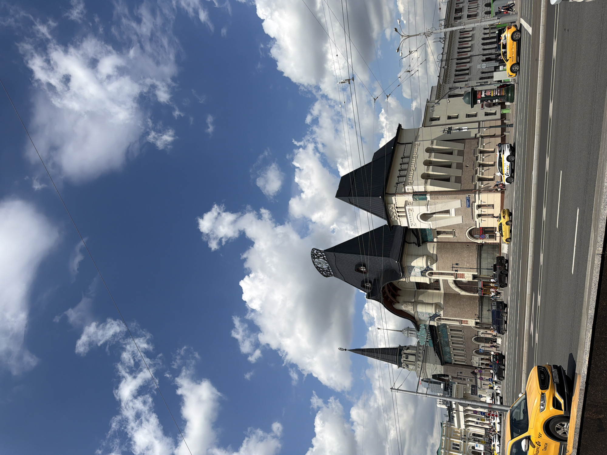 This is a ground-level, eye-level, slightly wide-angle outdoor shot of a street scene in front of a large, ornate building under a blue sky with fluffy white clouds. In the foreground, a yellow taxi cab is parked on the right side of the street. The street is asphalt with white lane markings and is busy with other vehicles, including more yellow and white taxi cabs, a dark blue minivan, and a black pickup truck. Pedestrians can be seen walking on the sidewalk in front of the building. The building itself is a grand structure with a mix of architectural styles, featuring dark, pointed roofs, light-colored walls with arched windows, and intricate decorative elements. The sky above is a vibrant blue, filled with cumulus clouds, and crisscrossed by overhead wires. The scene is brightly lit by daylight, suggesting it is daytime.