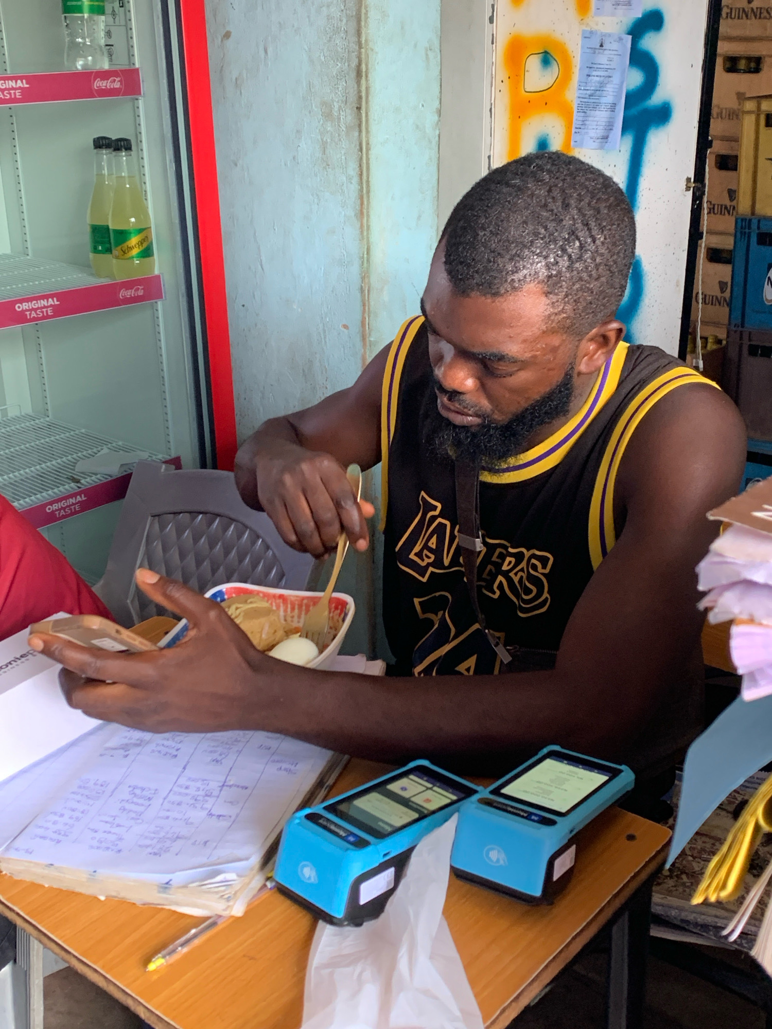 A man with dark skin and a beard is sitting at a wooden desk in what appears to be a small shop or convenience store, possibly in Jos, Nigeria. He is wearing a black sleeveless Lakers jersey with yellow trim. He is holding a smartphone in his left hand and a fork in his right hand, which is in a bowl of food containing what looks like eggs and noodles.
In front of him on the desk are two blue card readers, a thick notebook filled with handwritten entries, a pen, and a stack of papers. Behind him, there's a red-trimmed cooler displaying bottles of Schweppes and Coca-Cola. The wall behind the man is painted and has graffiti. There are also crates of Guinness beer stacked in the background. The lighting suggests it's daytime, possibly indoors with artificial lighting supplementing natural light. The overall scene depicts a transactional environment where the man might be managing his business while also taking a meal.