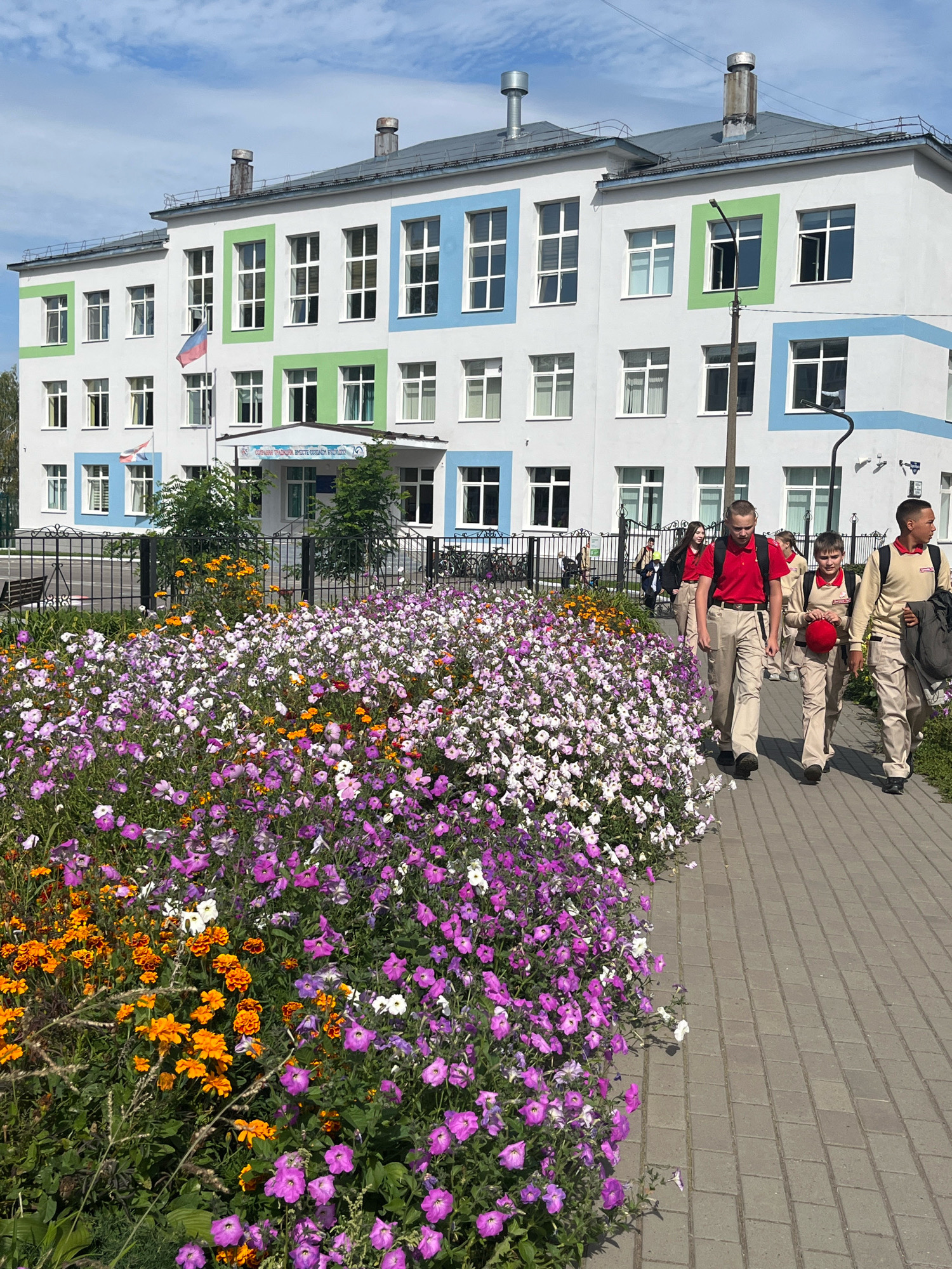 In Chaikovsky, Russia, a group of young people in school uniforms are walking on a paved sidewalk next to a vibrant flower bed. The school building, painted white with green and blue accents around its windows, stands in the background under a clear blue sky with scattered clouds. A Russian flag is visible flying on a flagpole in front of the school entrance. The flower bed is densely packed with purple, pink, and orange flowers, creating a colorful border along the path. The individuals are walking away from the camera, some carrying backpacks. The overall scene depicts a sunny day at a school with a well-maintained exterior.