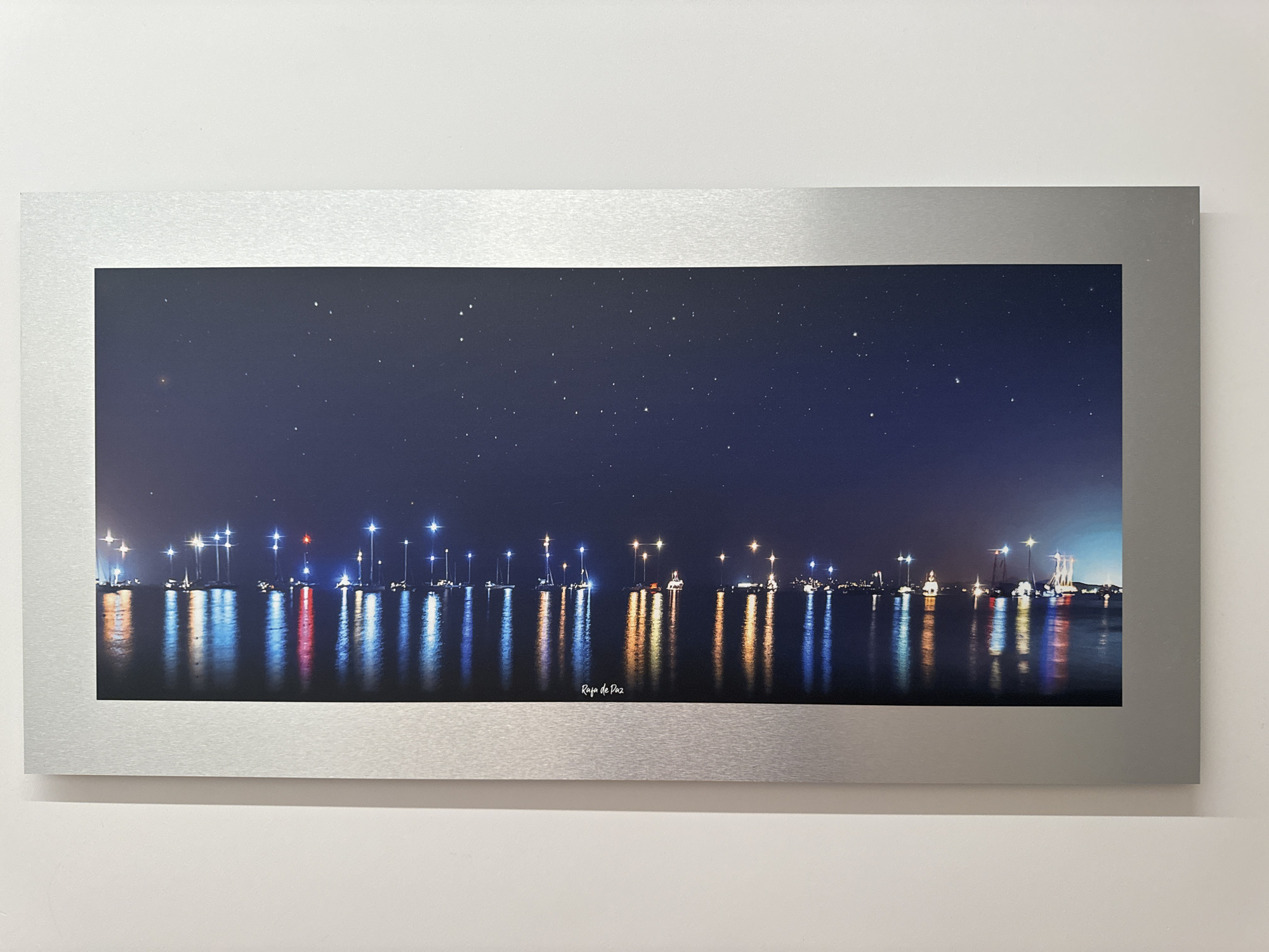The image displays a panoramic photograph of a nighttime harbor scene, likely in or near the city of Murcia, Spain, given the inscription "Rafa de Paz" which may be a signature or artist name, and the stylistic representation of a Mediterranean port. The photograph is framed by a brushed silver border and mounted on a white wall.
The photograph itself is dark, dominated by a deep navy blue sky filled with numerous small white stars. Below the sky is a body of water reflecting the lights from a distant shoreline. The shoreline is lined with numerous sailboats and other vessels, each illuminated by various lights. These lights create long, vibrant reflections on the water's surface, appearing as streaks of white, blue, red, orange, and yellow. Some of the boats have tall masts, creating distinct vertical lines of light against the dark water. The overall impression is of a calm, star-filled night in a bustling harbor.