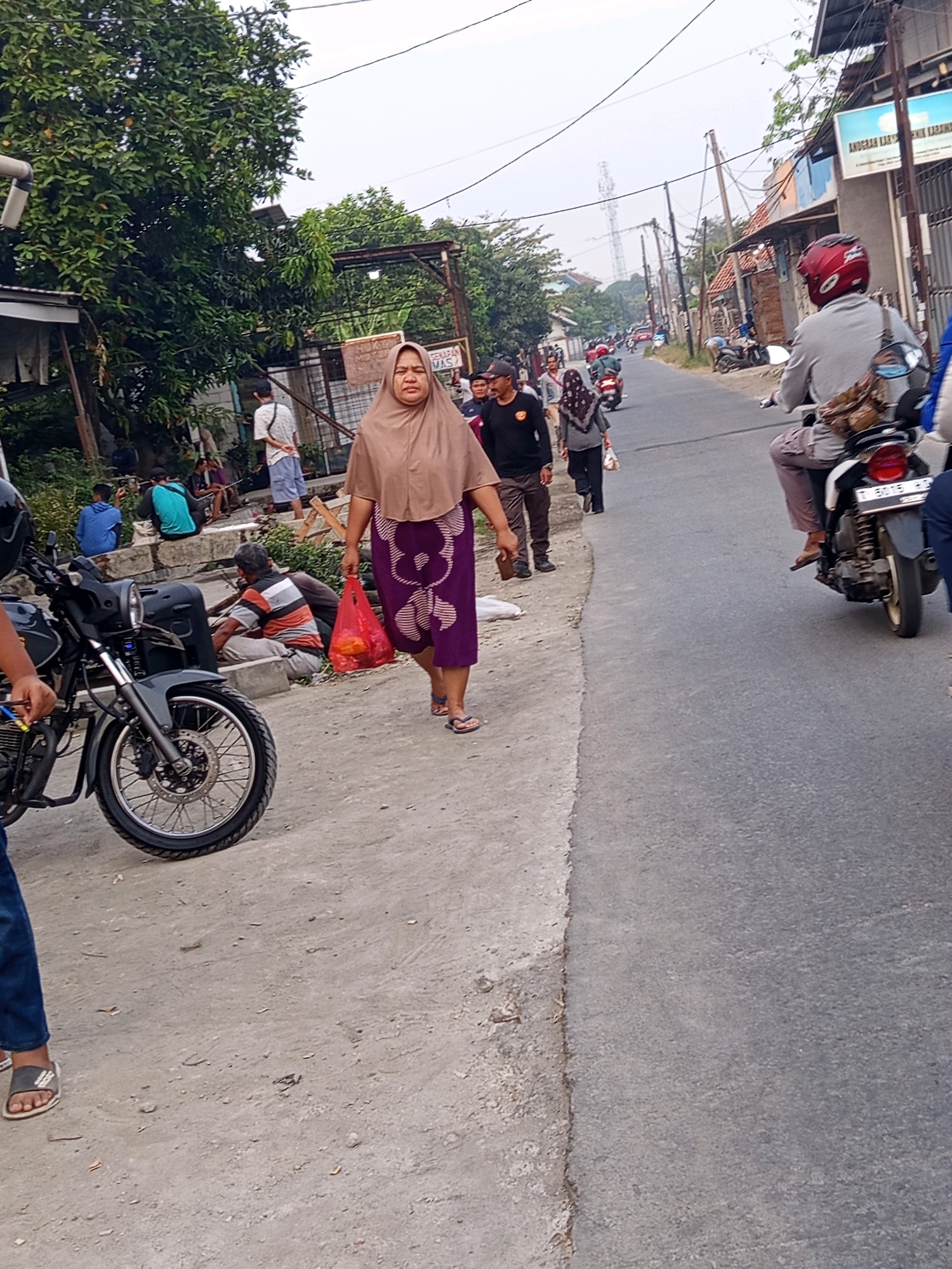 The image shows a street scene in Karawang, Indonesia, with a focus on several individuals and vehicles. In the foreground, a woman wearing a hijab and a patterned purple skirt walks towards the right, carrying a red plastic bag filled with items. Next to her, to the left, a motorcycle is parked, with its front wheel and handlebars visible. Further down the street, other people are seen walking and sitting. Some are on motorcycles, suggesting traffic is present. The environment appears to be a typical urban or suburban street, with buildings and trees lining the road. The sky is overcast, indicating a daytime scene.