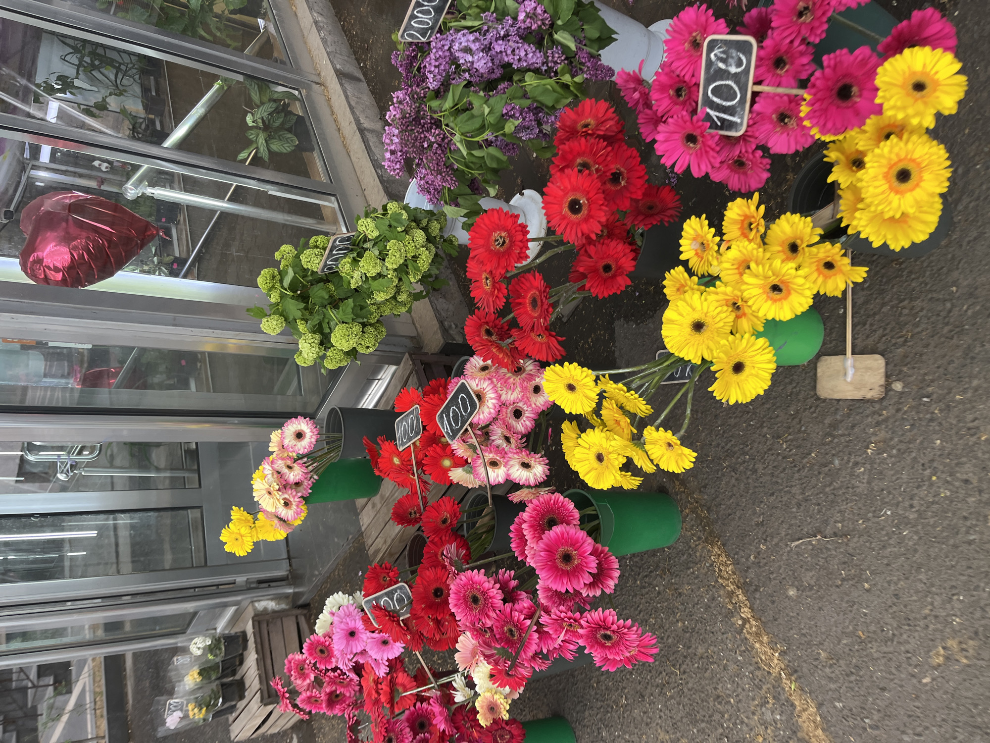 The image shows a flower stand outside a store, likely in Yerevan, Armenia. There are several buckets of flowers for sale including purple lilacs, lime green hydrangeas, red, pink and yellow gerbera daisies. Small black signs indicate a price of 100 or 2000 for the flowers. A red heart-shaped balloon is visible inside the store. The stand is located on a sidewalk or pavement area.