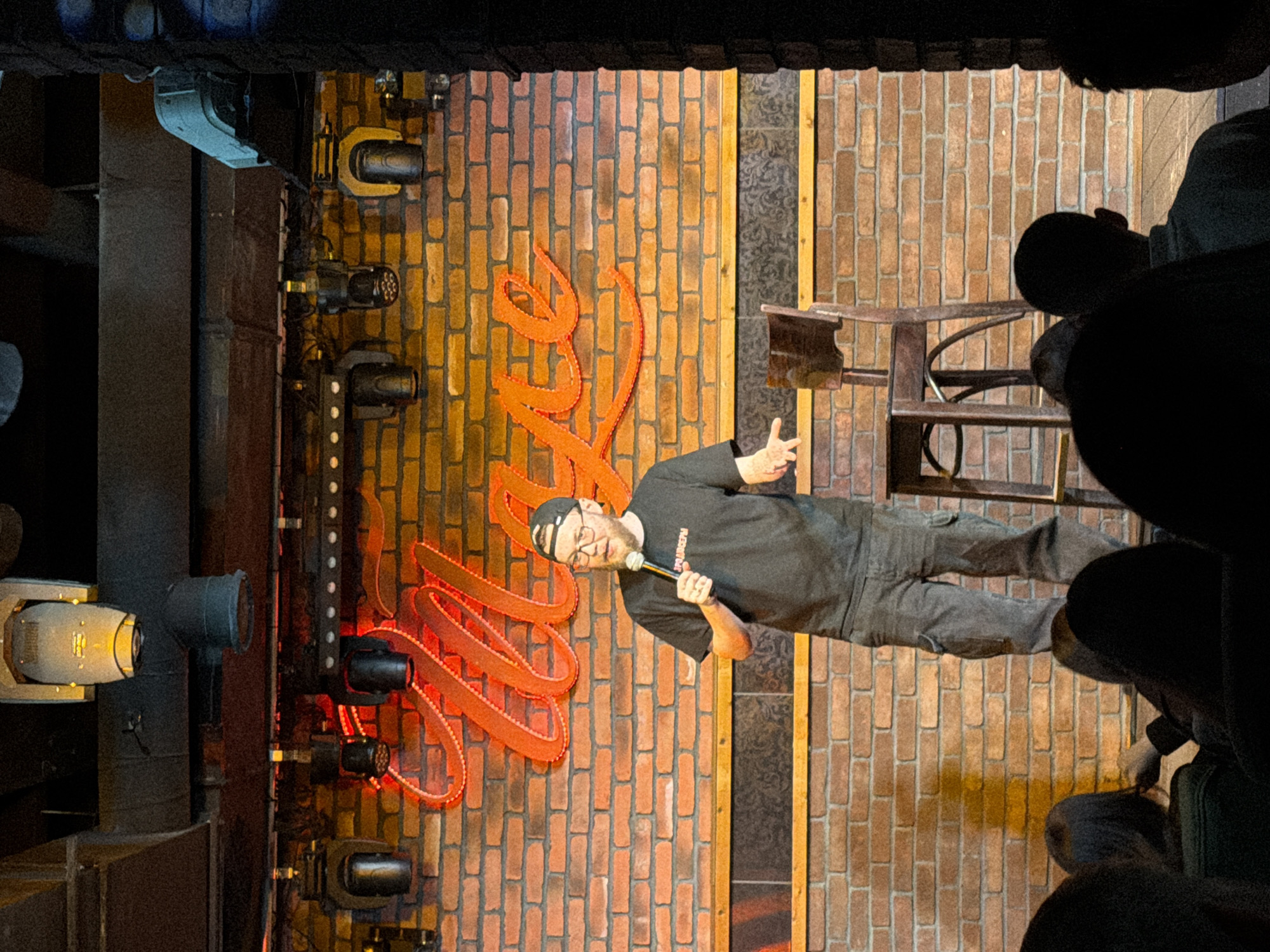 A man on stage is performing stand-up comedy in Klin, Russia. The man is wearing a black t-shirt and dark pants, holding a microphone. The stage has a brick wall backdrop with orange neon signs. There is stage lighting to the left and an audience to the right. A wooden stool is visible in the background.