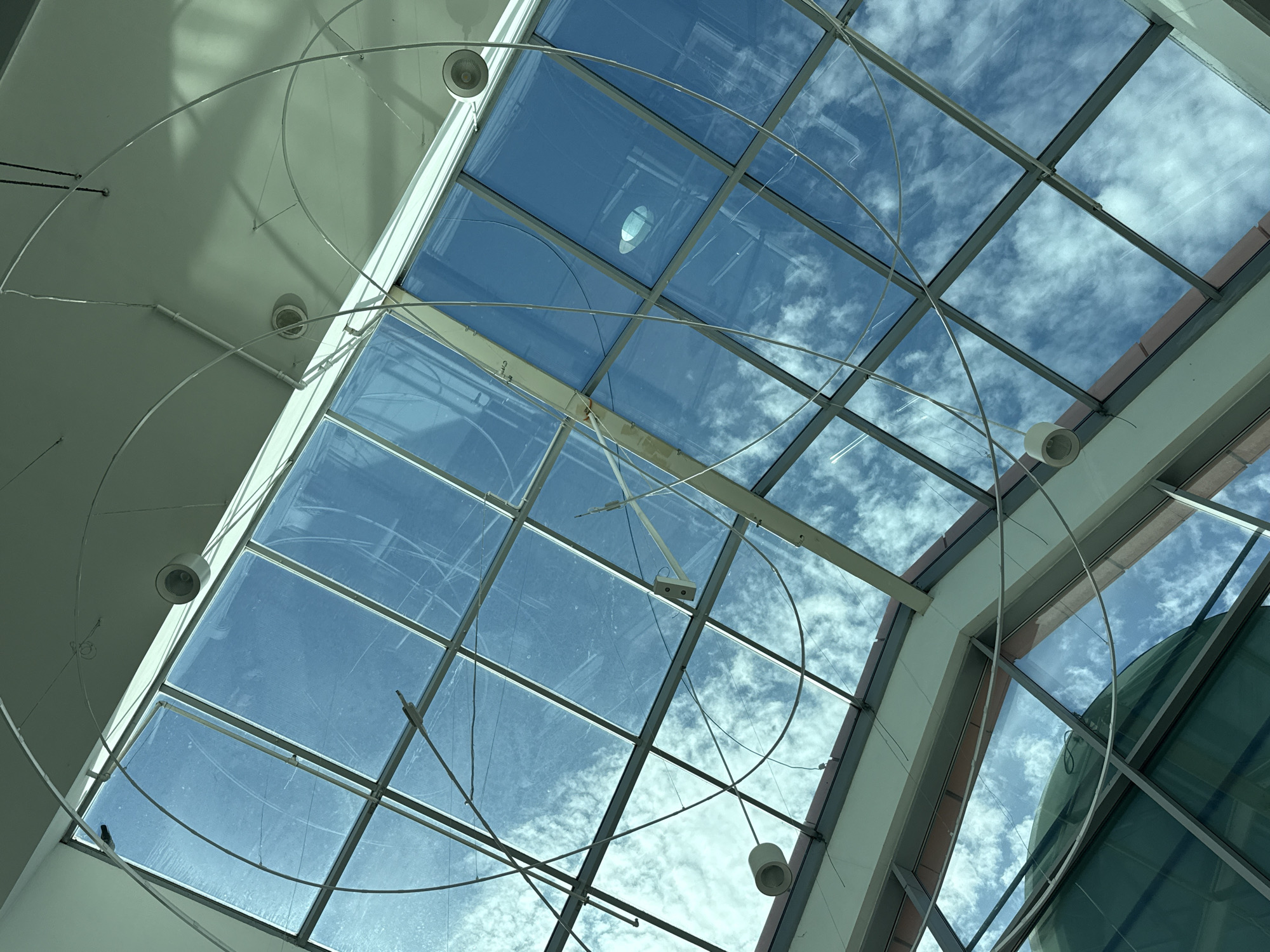 The image shows a view looking upwards towards a glass-paneled ceiling, likely a skylight, in a building. The glass panes are crisscrossed by metal framing and suspended by wires are several circular light fixtures. The sky is visible through the glass, with scattered white clouds against a bright blue background. Part of the building's interior structure, including the white ceiling and some metallic supports, is also visible. The location is identified as Bielany Wrocławskie, Poland.