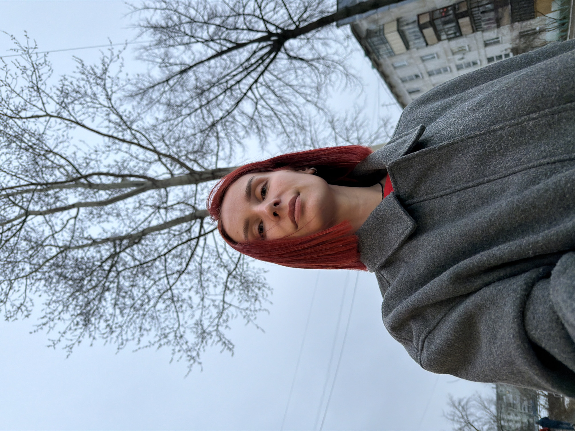 A person with short, red hair, wearing a gray coat, is the focal point of the photograph. The image is taken from a low angle, looking up at the individual. The background consists of a leafless tree with numerous branches against a cloudy sky. In the upper right corner, a multi-story building is visible. The location is Chaikovsky, Russia. There are power lines.