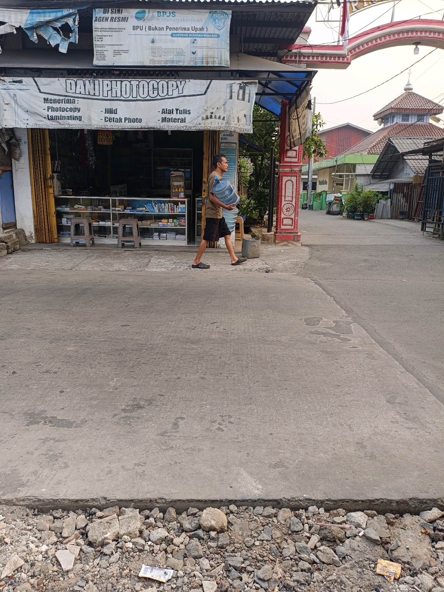 A man walks on a concrete sidewalk carrying a large blue water jug. He is wearing a brown t-shirt and black shorts. The man is walking past a shop with a yellow awning and glass display cases filled with merchandise. A banner above the shop reads "DANI PHOTOCOPY". In the background, a street leads towards buildings and a mosque with a dome and minaret. The scene appears to be in Tangerang, Indonesia, based on the signage and architectural style. The foreground is dominated by a rough, broken concrete area with scattered rocks and debris.