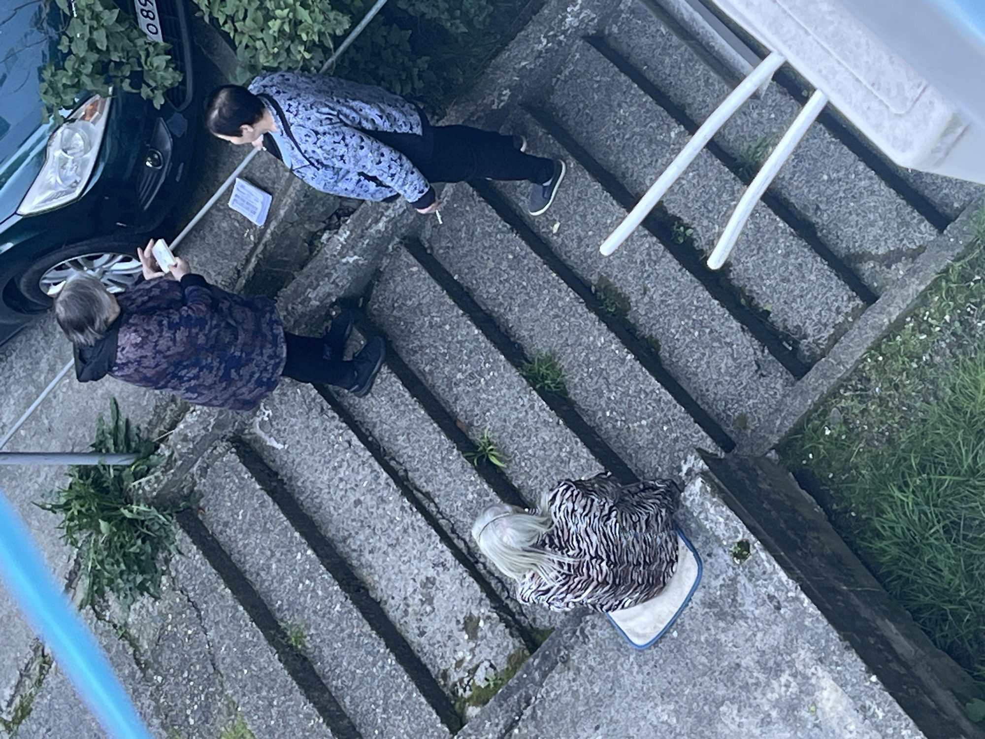 An overhead shot captures an outdoor scene featuring three individuals on a set of grey concrete stairs, with lush green vegetation and a partially visible car to the left. The location is contextualized as Sochi, Russia.
In the upper-left, a dark-colored car, possibly green or dark blue, shows its front wheel and headlight, with a partial license plate displaying "580". Dense green foliage borders the car.
Two individuals are standing on the concrete stairs. The person in the upper part of the frame wears a light blue/grey patterned jacket and dark pants, with short dark hair. They appear to be holding a lit cigarette in their right hand. A small white piece of paper or card lies on the step near their feet. Slightly below and to the left, another person, with light grey hair tied back, wears a dark patterned jacket and dark pants. This individual is holding a white smartphone with both hands, possibly taking a picture, video, or observing its screen. Both standing individuals are looking downwards, seemingly focused on something on the ground or the third person.
The third individual is positioned further down the stairs, lying on their side on a light-colored mat or cushion with a blue trim. This person wears a black and white striped or patterned top and has long blonde or white hair. Their posture suggests they are either resting or in some form of distress, though emotions are not clearly discernible from this angle.
White railings or bars are visible on the right side of the frame, possibly indicating a balcony or a part of the stair structure. Patches of grass and small weeds grow between the concrete steps and along the edges of the stairs, particularly on the right side. The lighting suggests it is daytime, possibly overcast or during the softer light of early morning or late afternoon. The overall scene depicts an everyday outdoor environment with an unusual concentration of focus on the person lying down.