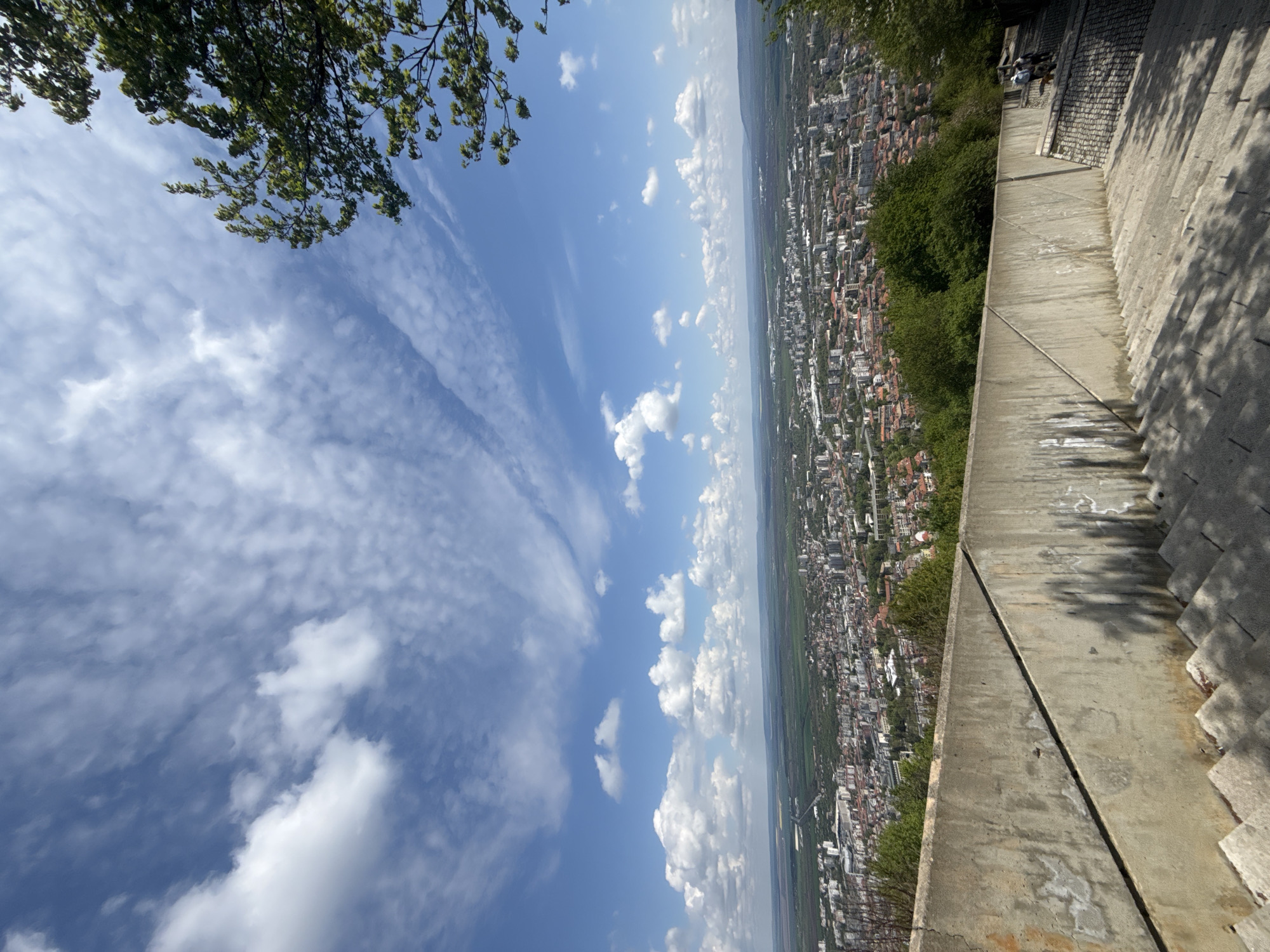 The image presents a sweeping panoramic view of a city sprawling across a landscape, captured from a high vantage point under a bright, sunny sky. The perspective suggests the photographer is standing on an elevated platform or hillside, looking down and out over the urban area.
The top and left portions of the frame are dominated by a clear blue sky, adorned with scattered white clouds of varying types—some appear as fluffy cumulus, while others are stretched into wispy, almost cirrus-like formations. A cluster of vibrant green tree branches and foliage extends into the upper left corner, partially framing the sky.
On the right side of the image, a robust concrete structure occupies the immediate foreground. This appears to be a wide walkway, terrace, or part of a larger monument, featuring visible steps on its far right edge and distinct shadow patterns. Several small human figures are discernible on this concrete structure towards the upper right, suggesting people are present, perhaps observing the view.
The vast cityscape of Shumen, Bulgaria, unfolds across the middle and lower-left sections of the image. It comprises a dense collection of buildings, many characterized by red and orange tiled roofs, interspersed with patches of green vegetation and trees. The urban development extends far into the distance, eventually meeting a distant horizon line where the land flattens into what appears to be a plain or valley.
The bright sunlight indicates it is daytime, likely late morning or early afternoon, with clear weather. There is no visible text in the image. The scene conveys a sense of grandeur and scale, showcasing the expansive urban environment juxtaposed with natural elements from a commanding elevated perspective.